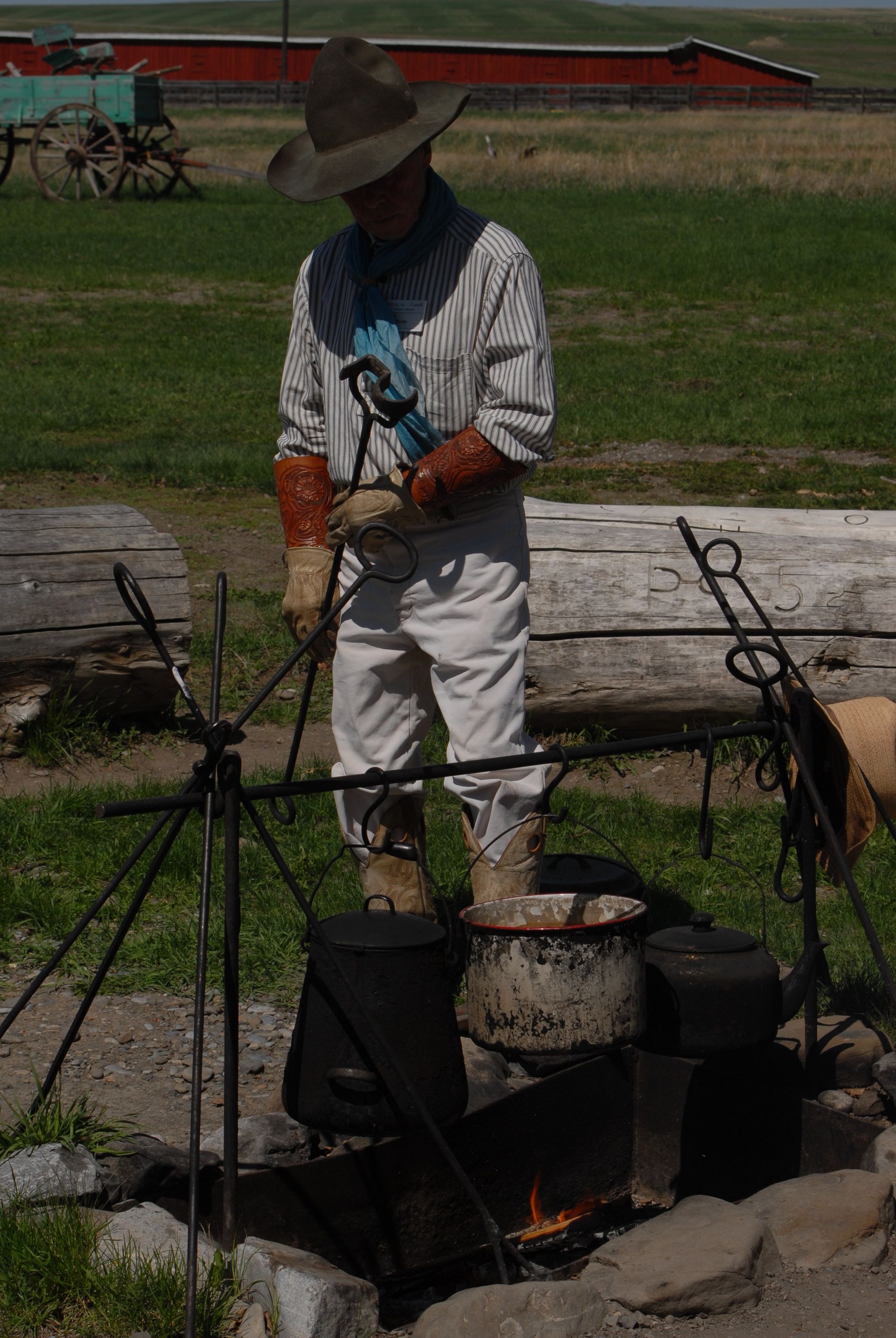 Cowboy tending a fire, cooking food in a pot, standing outdoors near a wooden wagon and red barn.