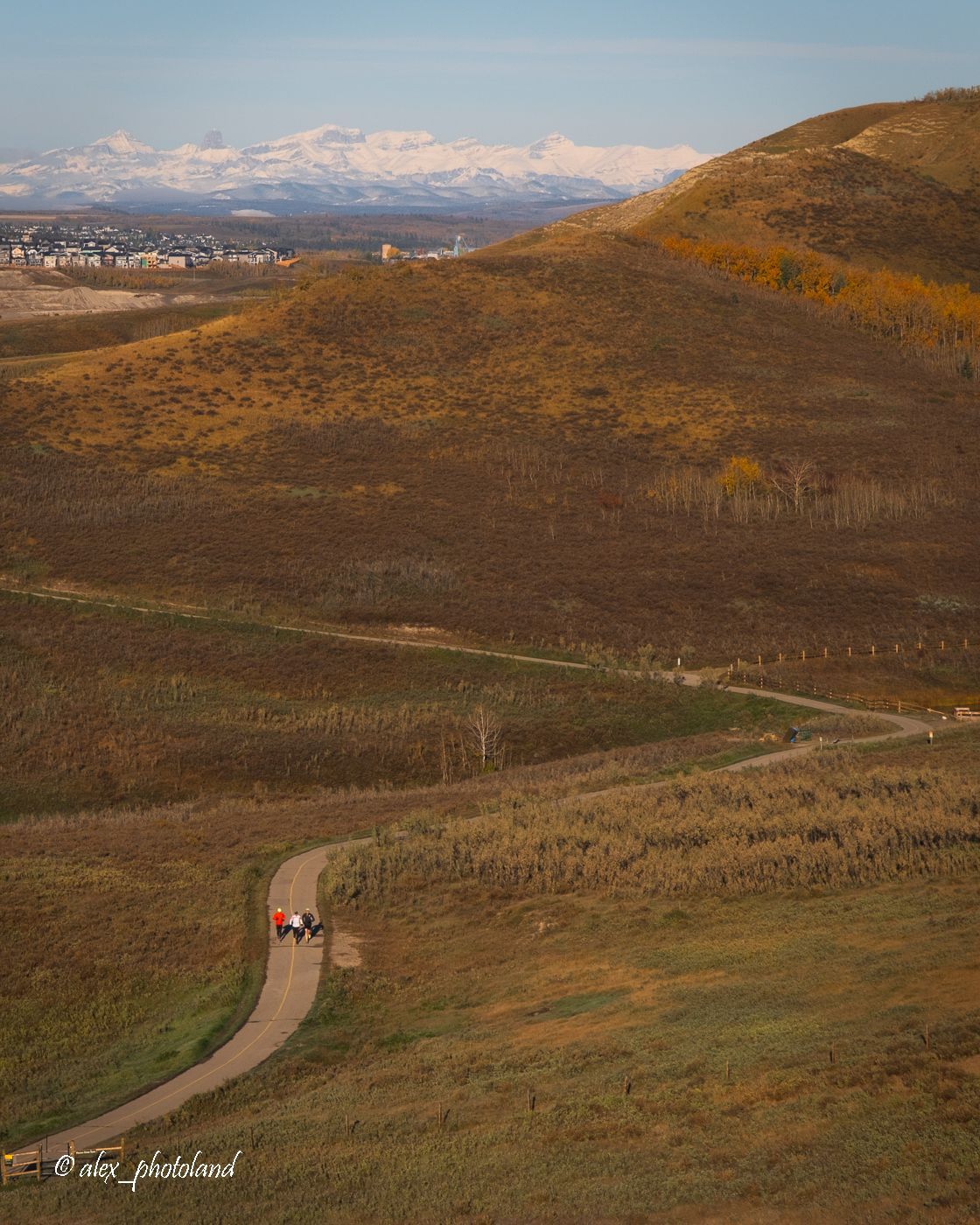 Rolling hills with a winding path and a few hikers. Snowy mountains and town in the distance.