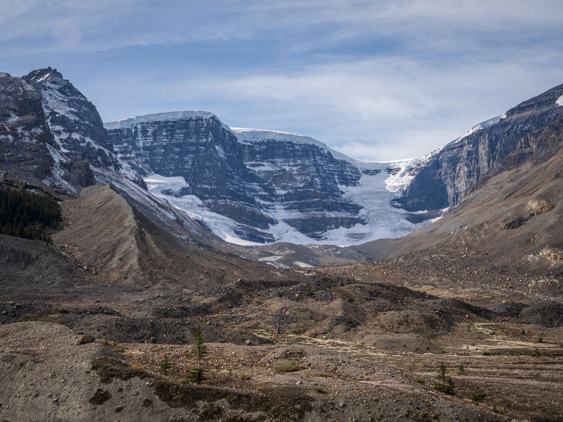 Snow-covered mountain range with glacier in a valley. Brown rocky terrain in the foreground under a blue sky.
