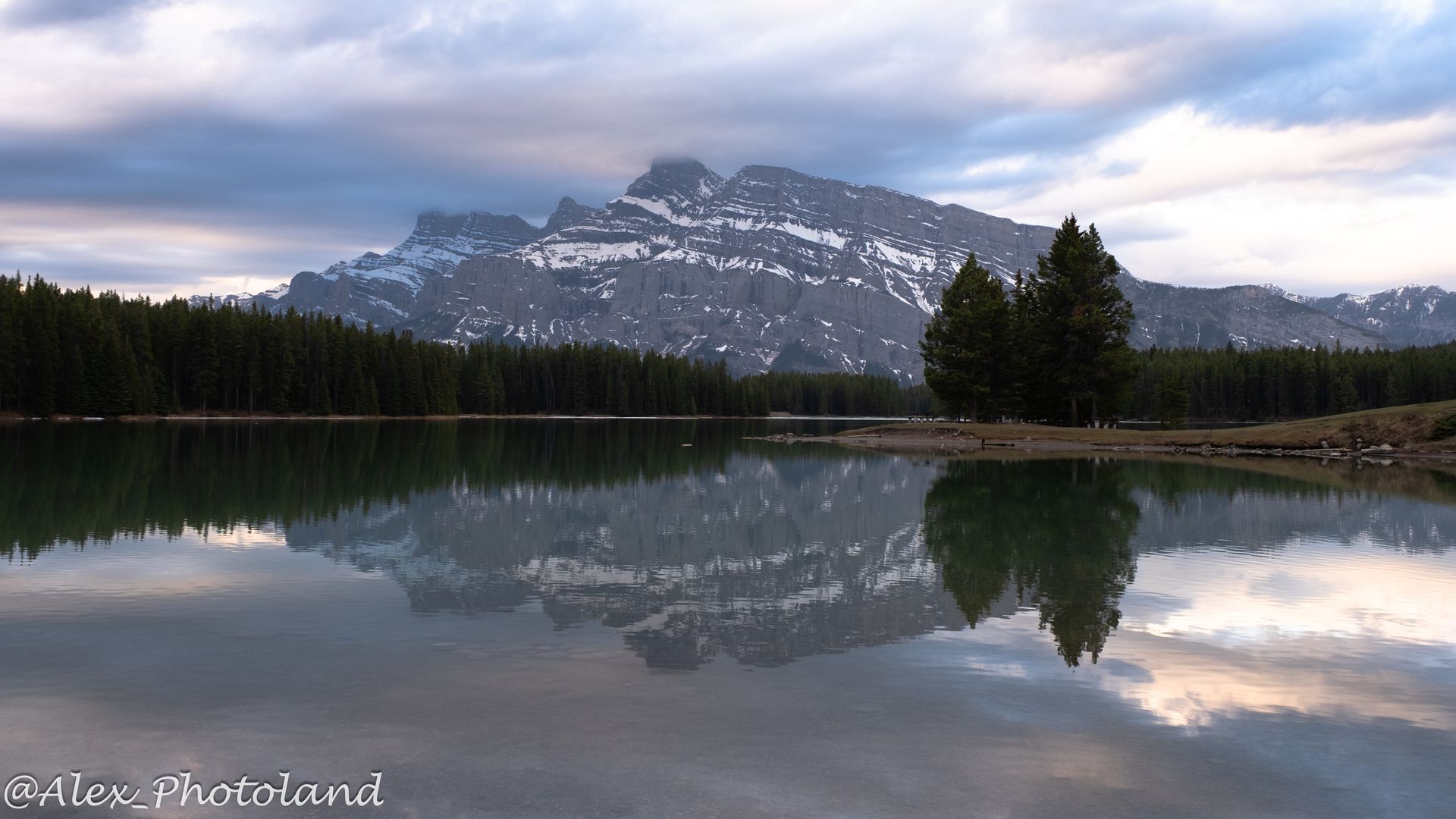Lake reflecting a snow-capped mountain under a cloudy sky, with evergreen trees along the shoreline.