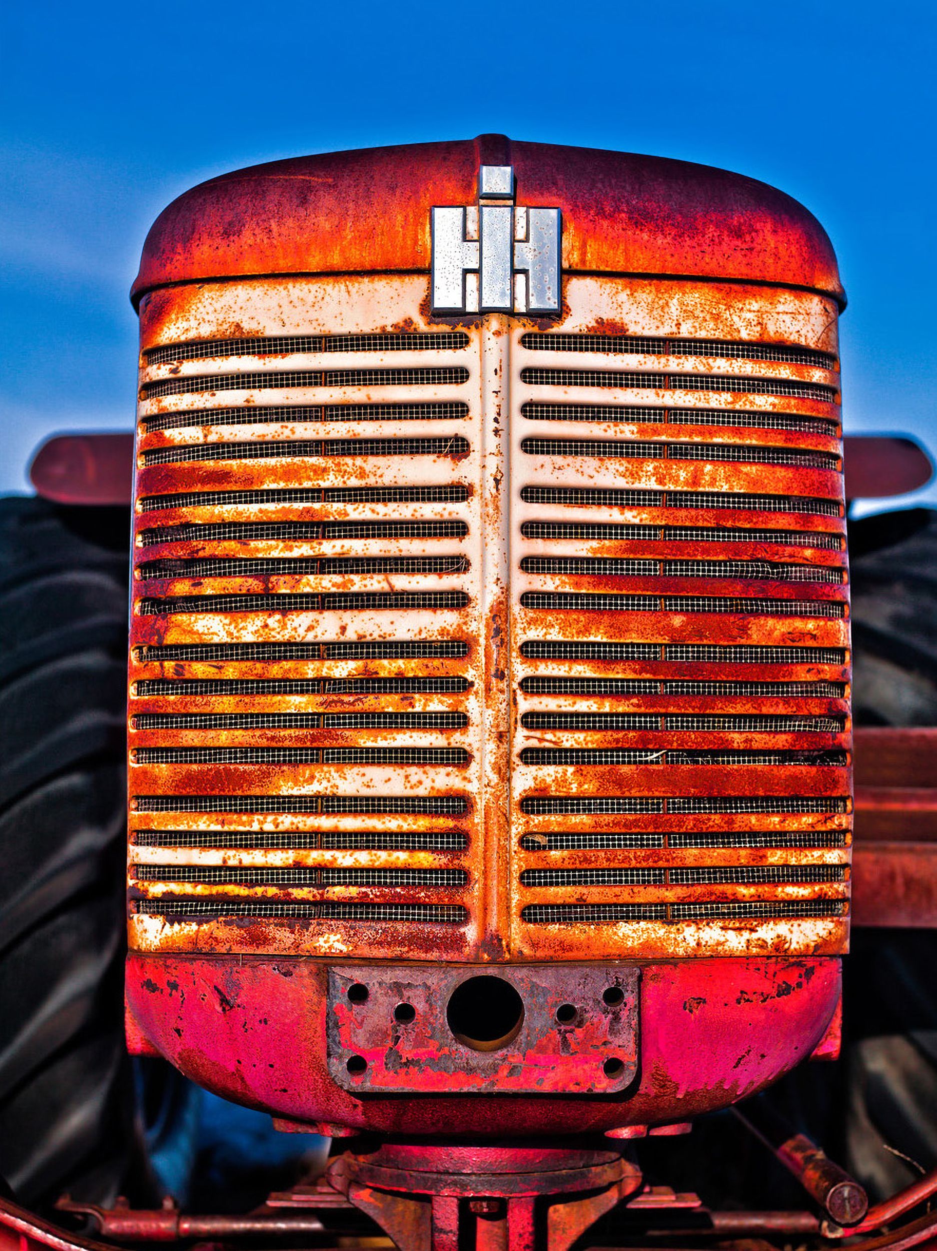Rusty red International Harvester tractor grill with silver logo, against blue sky and tractor tires.