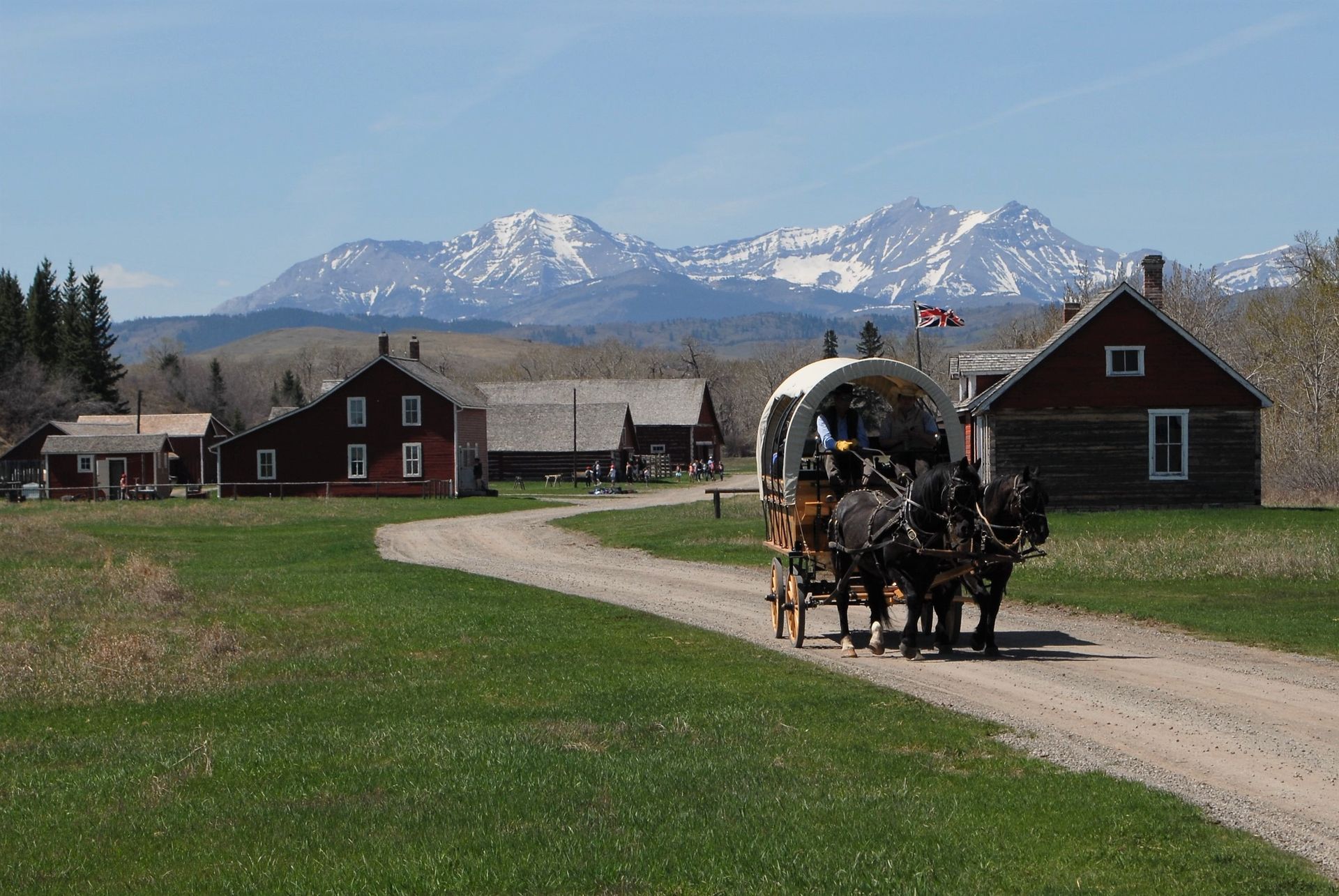 Covered wagon pulled by horses on dirt road, passing buildings and mountains.