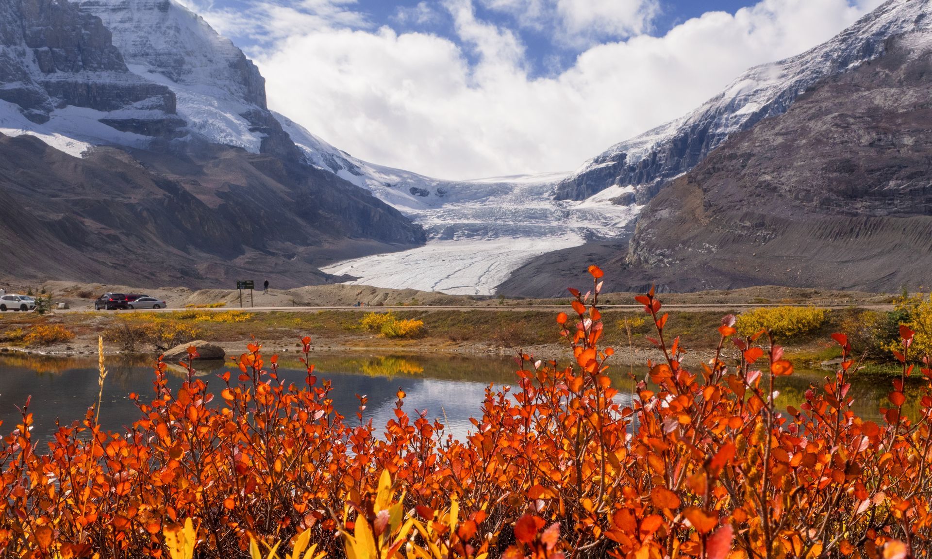 Autumn landscape with red foliage, lake, road, and a glacier between snow-capped mountains.