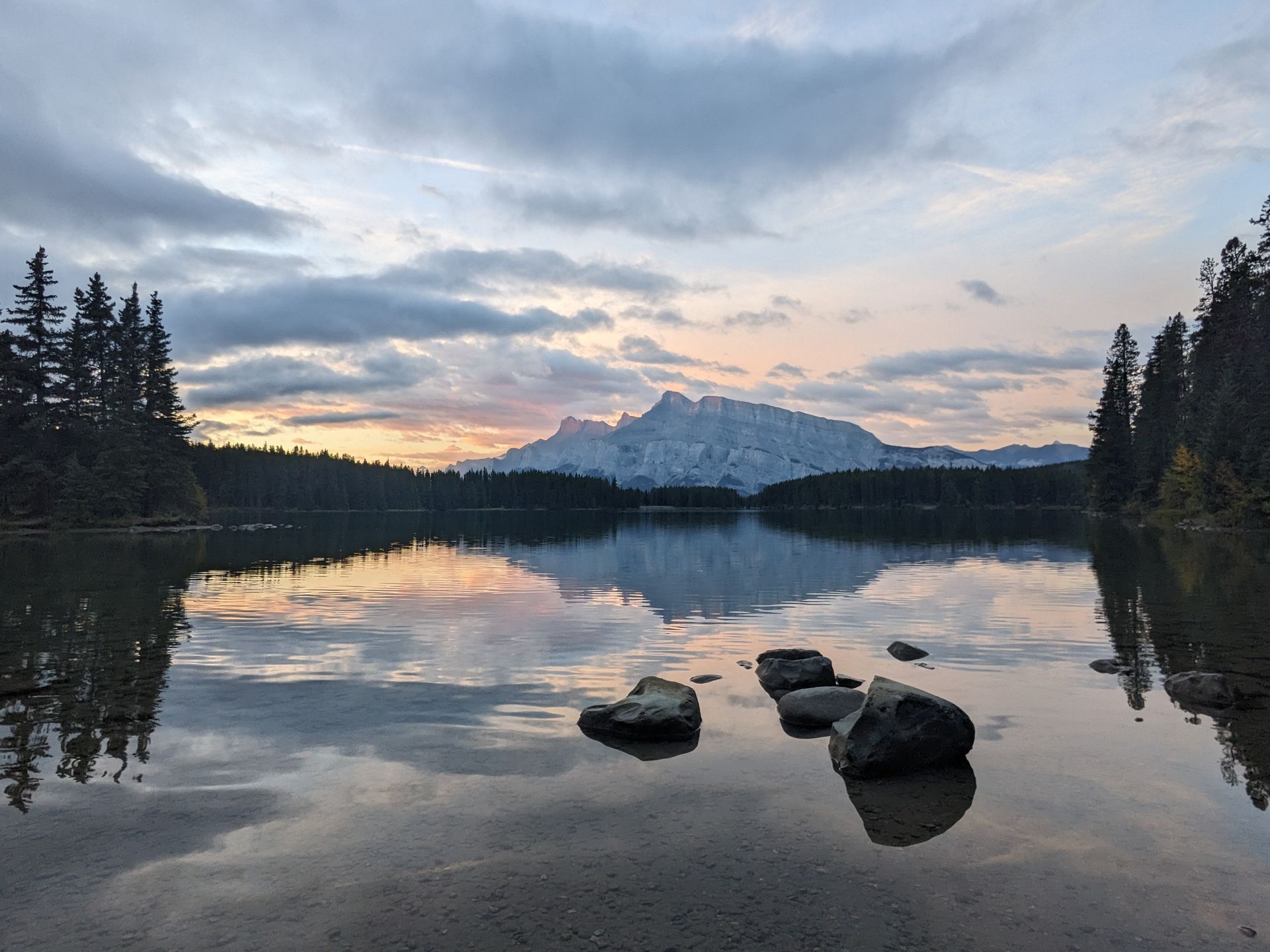 Lake with mountain reflection under a cloudy sky at dusk. Rocks in foreground.