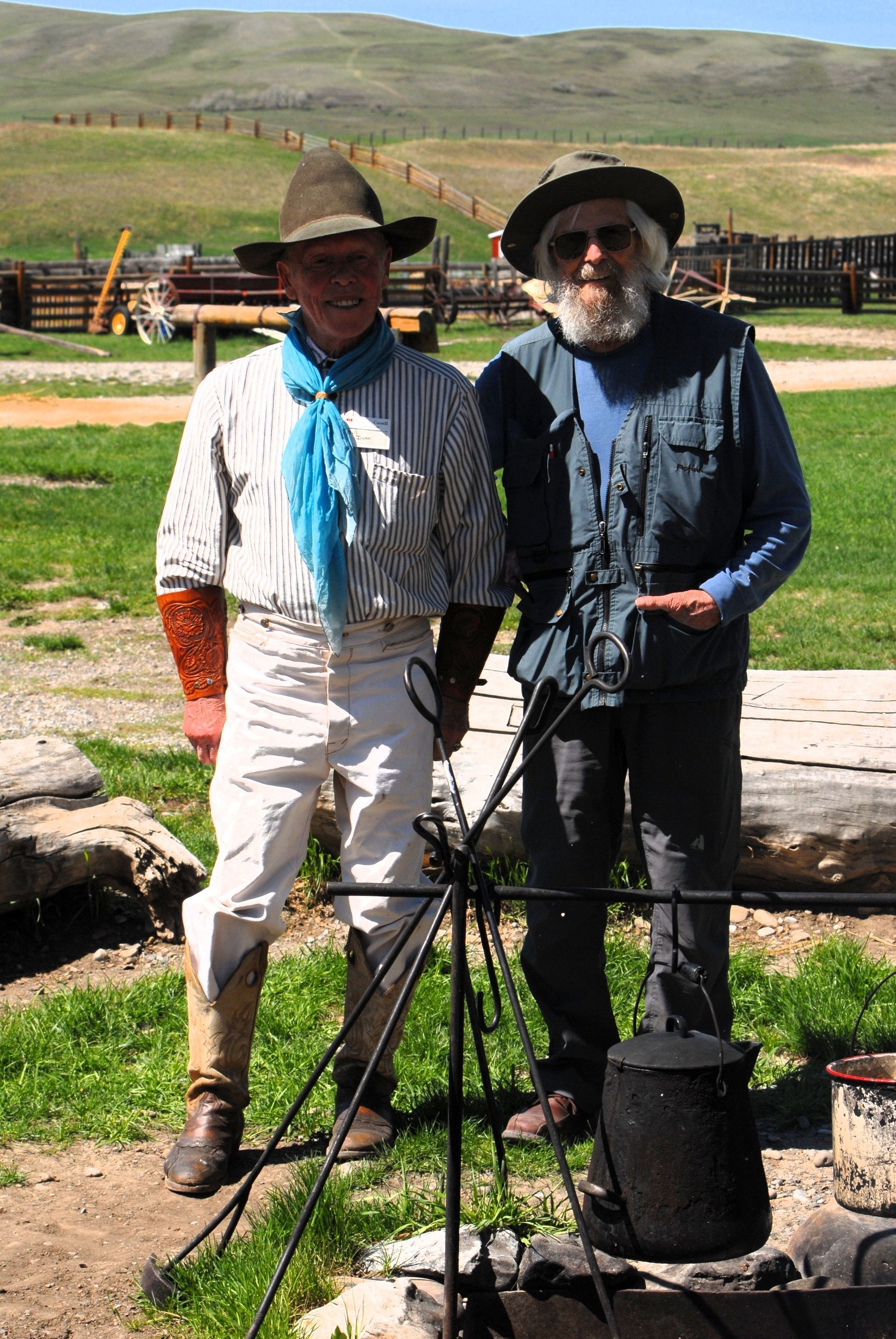 Two men in cowboy attire standing near a cooking pot outdoors.