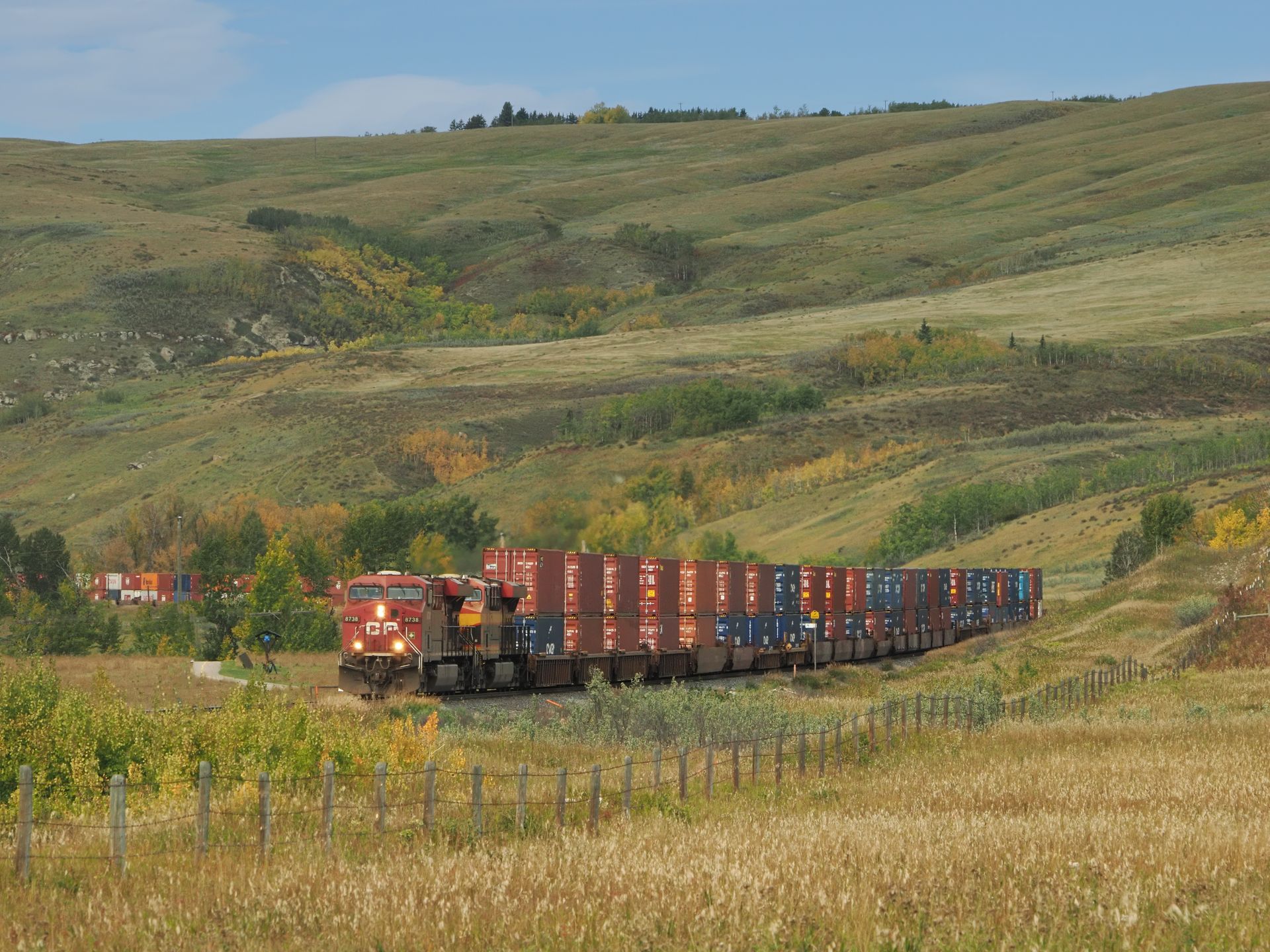 Train hauling shipping containers through a grassy landscape with rolling hills under a blue sky.
