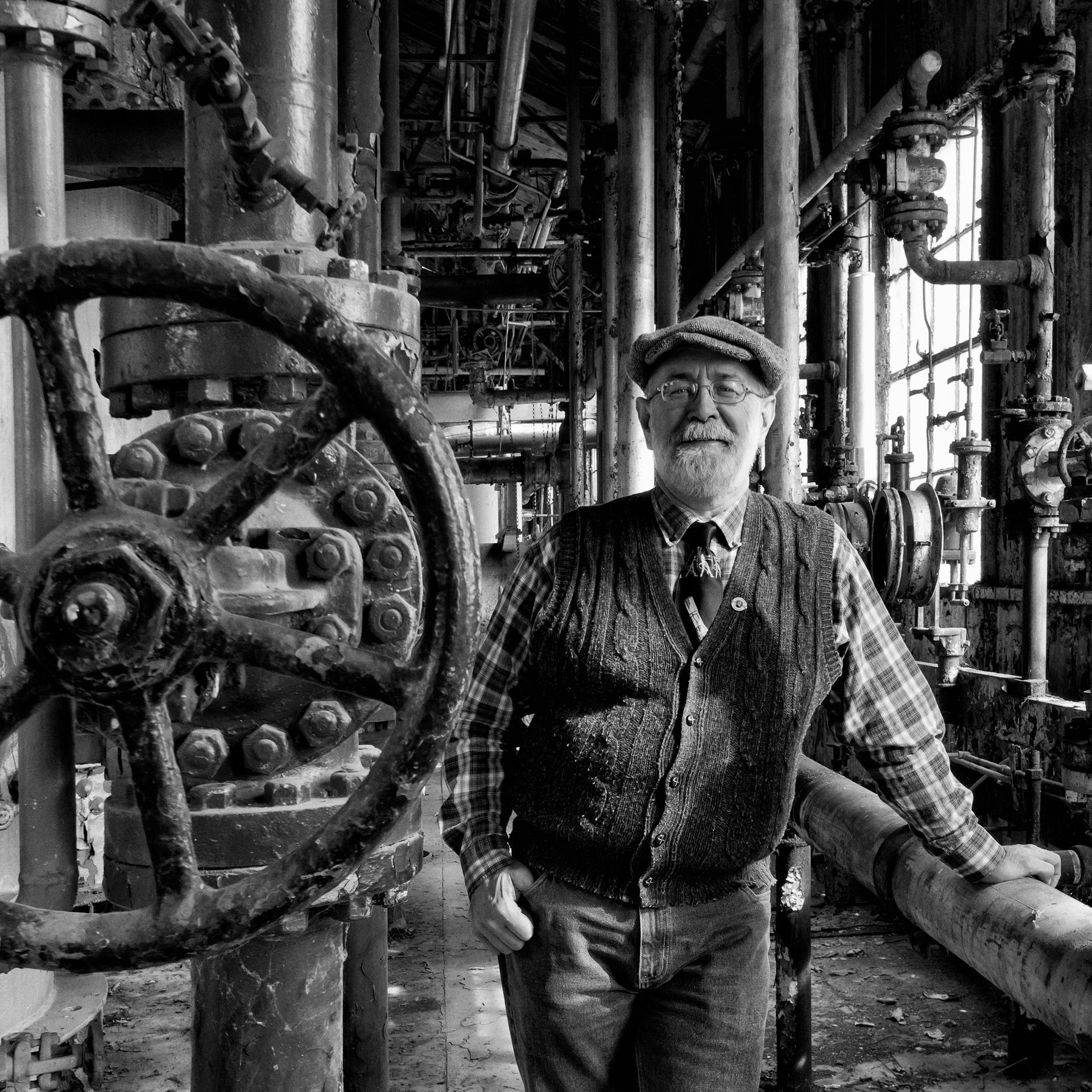 Man in work clothes and cap, smiling, leans on pipes with large valve in industrial setting.