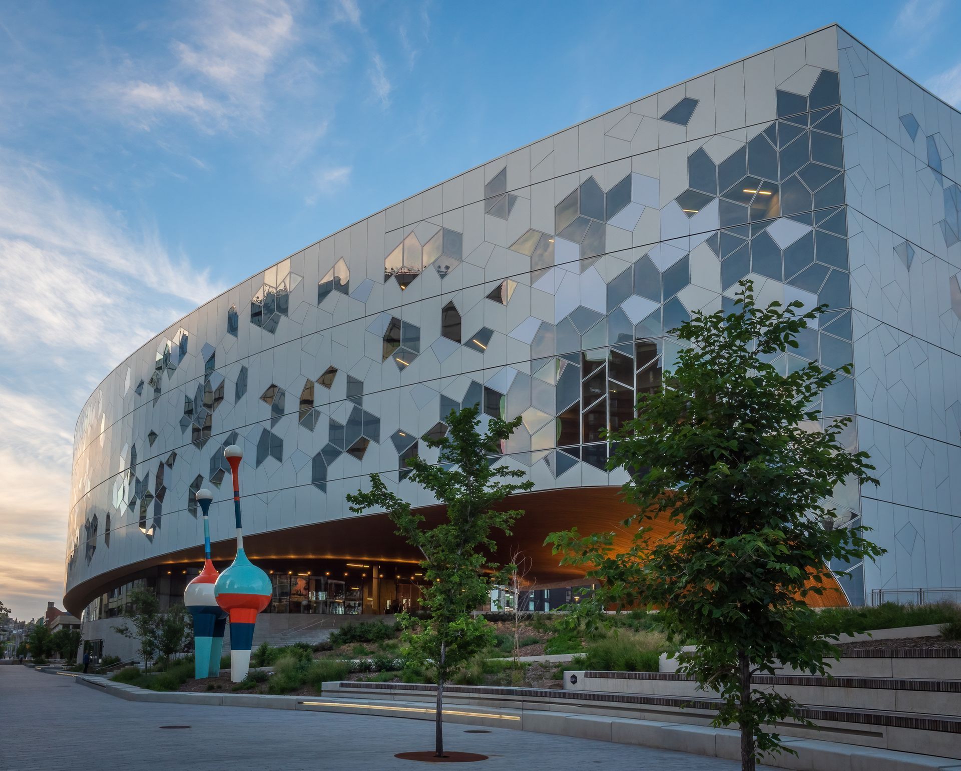 Modern, curved building with geometric facade. Trees and colorful sculptures in front, blue sky in background.