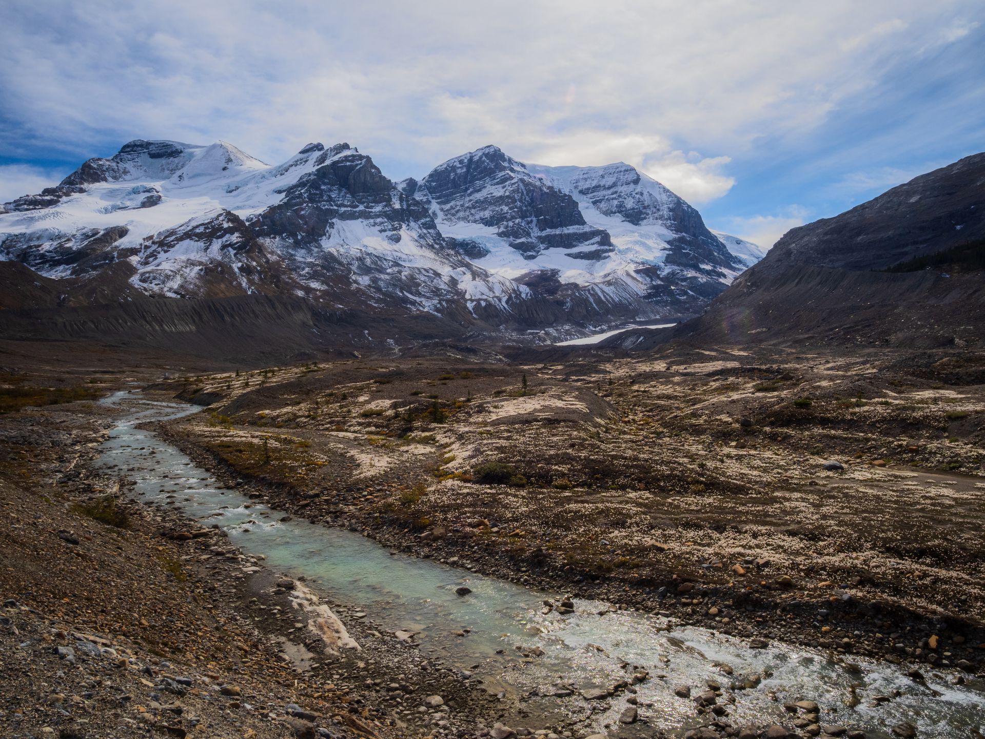 Snow-capped mountains tower above a glacial valley with a river winding through rocky terrain under a cloudy sky.