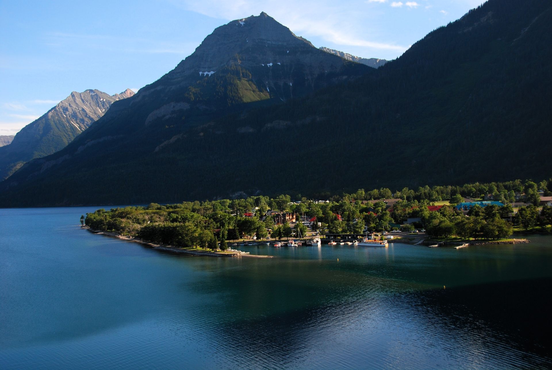 Lakeside village at the foot of mountain range. Blue water reflects mountains, trees, and buildings.
