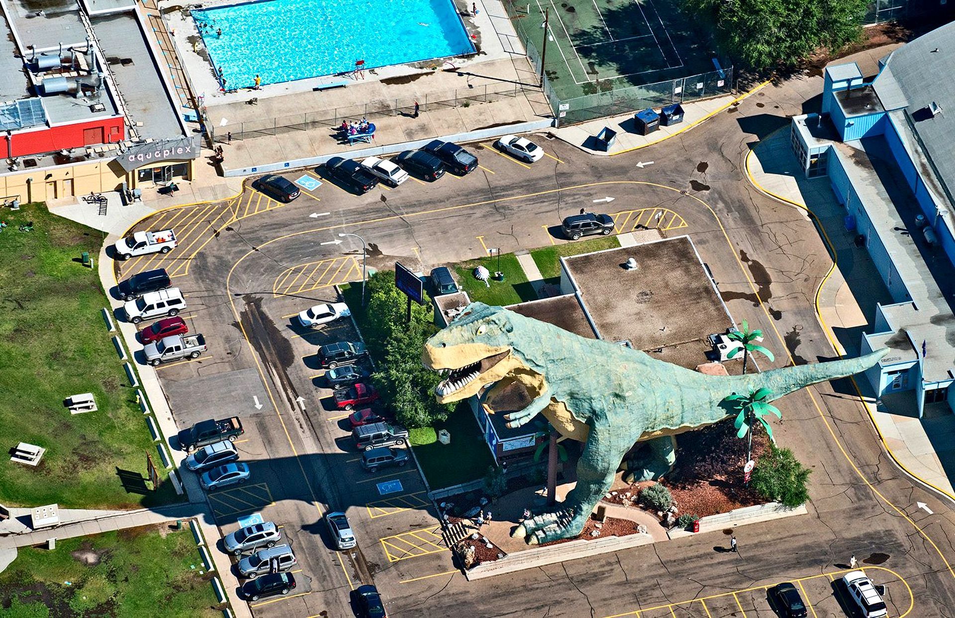 Aerial view of a large green dinosaur statue in a parking lot, near a swimming pool.
