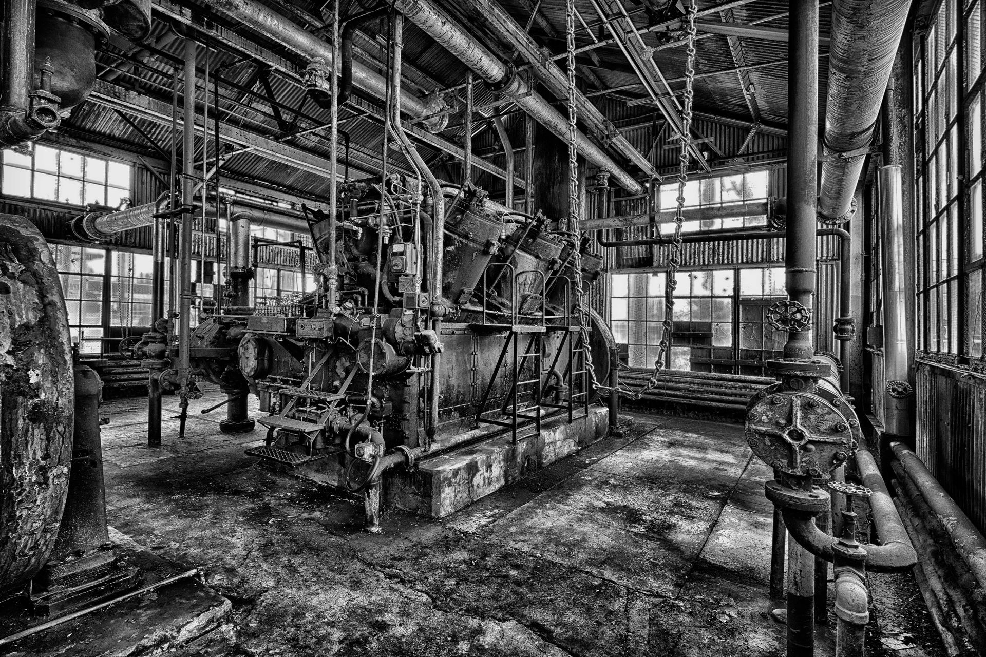 Black and white shot of an abandoned industrial interior, machinery and pipes overgrown with rust.