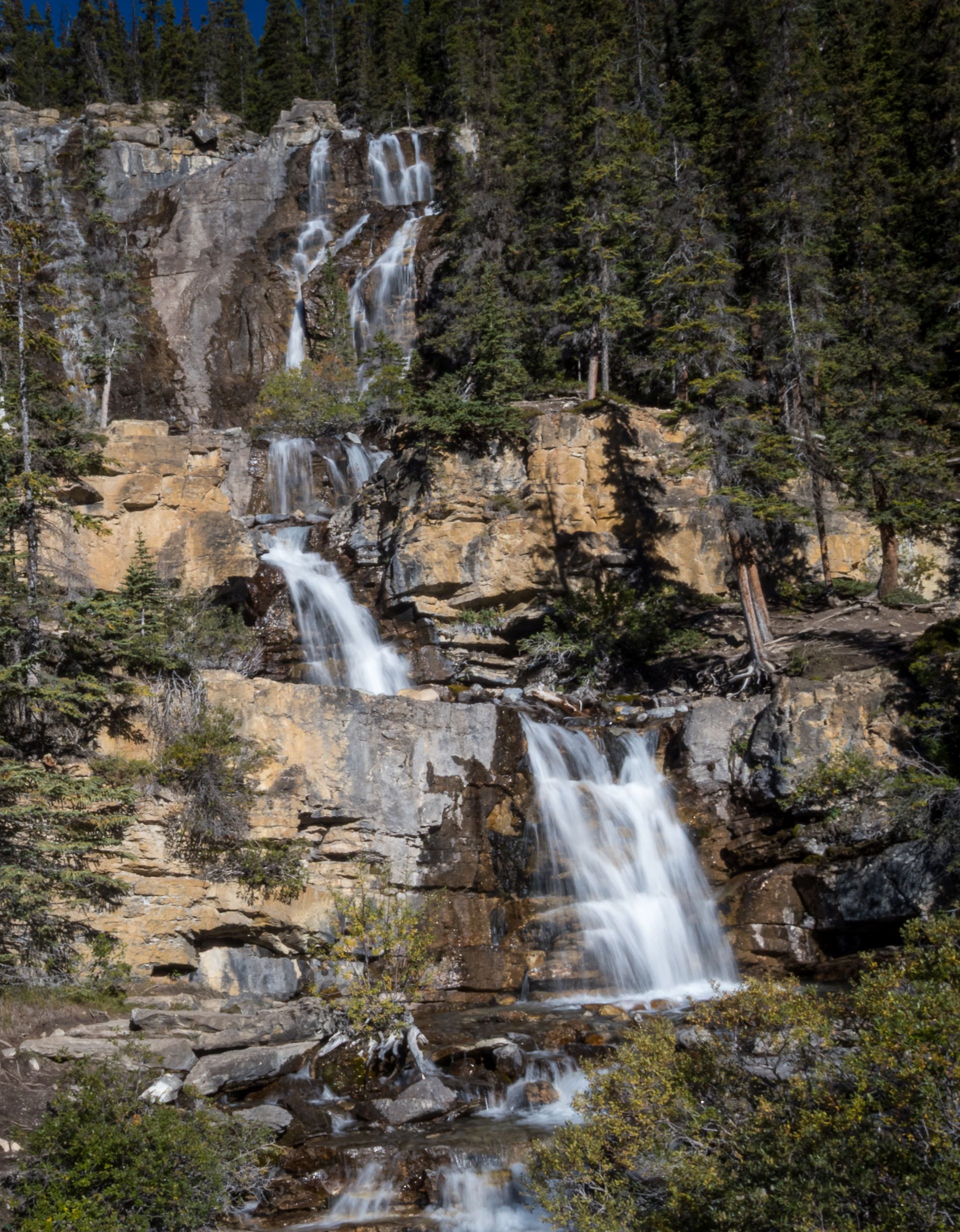 Waterfall cascading down a rocky cliff face, surrounded by green trees in a forest.
