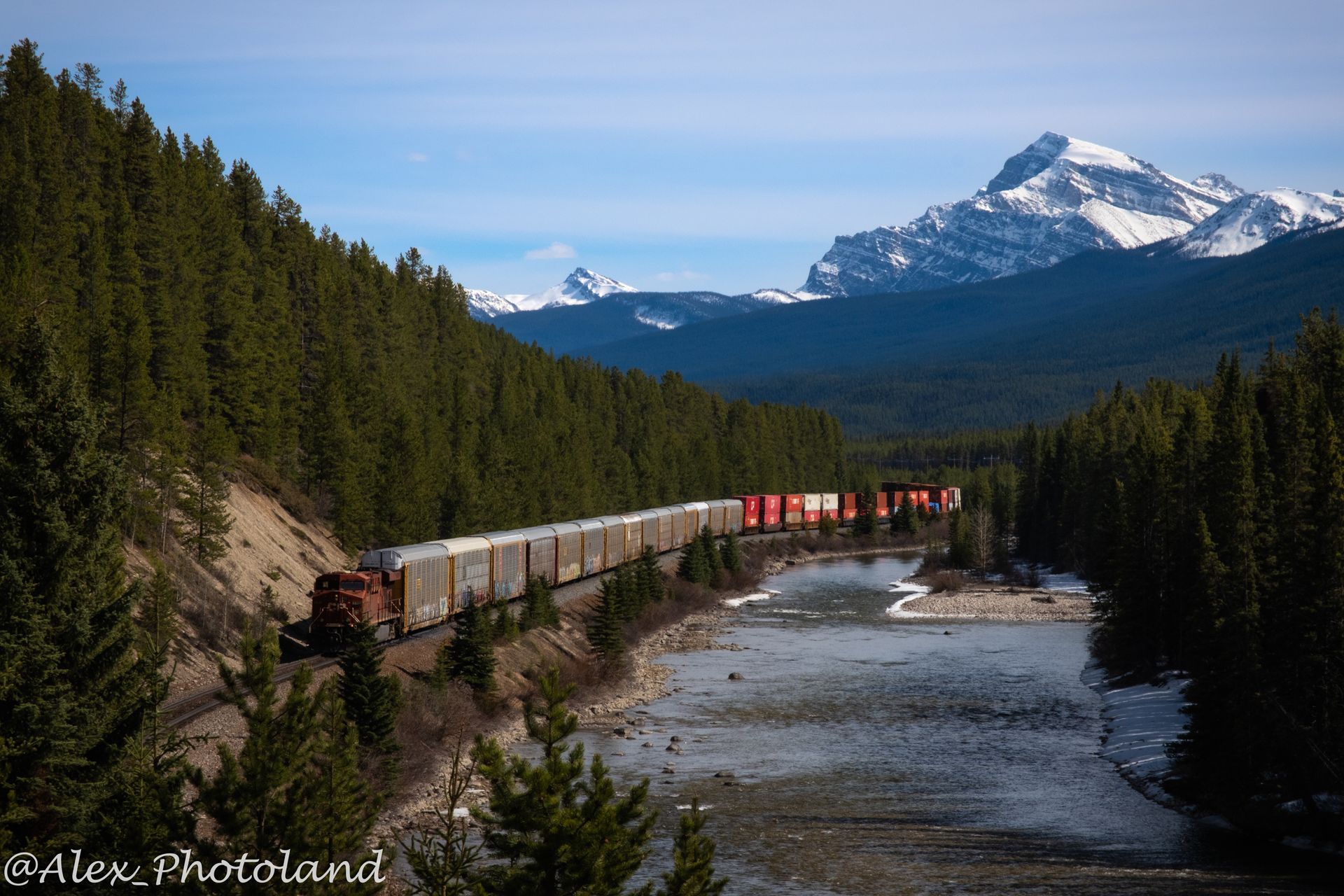 Train travels along river through forested valley, snow-capped mountains in the distance under a blue sky.