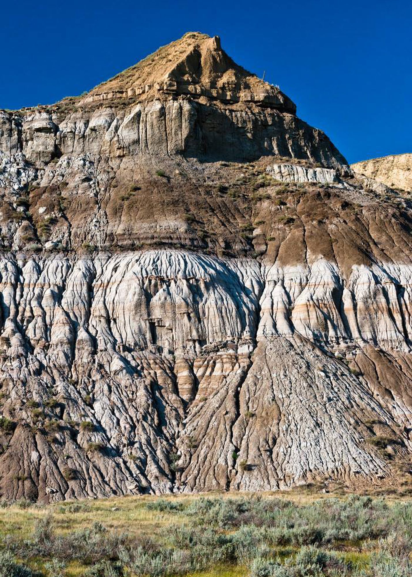 Layered mountain in shades of brown, white, and tan against a blue sky, with grassy foreground.