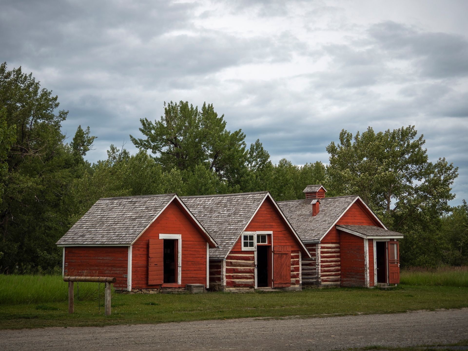 Red wooden sheds with open doors sit on a grassy area, trees in the background, cloudy sky.