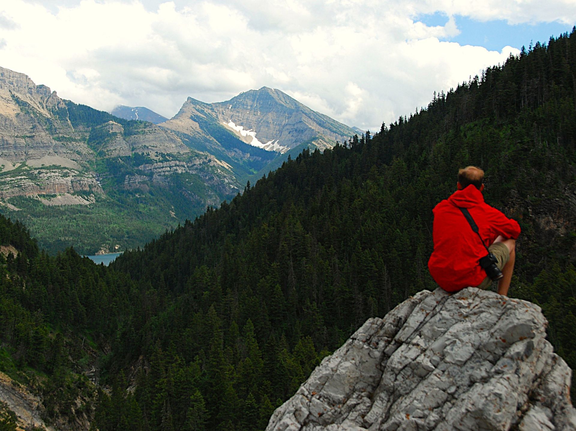 Person in red jacket sits on rock, overlooking a mountain valley with forest and lake.