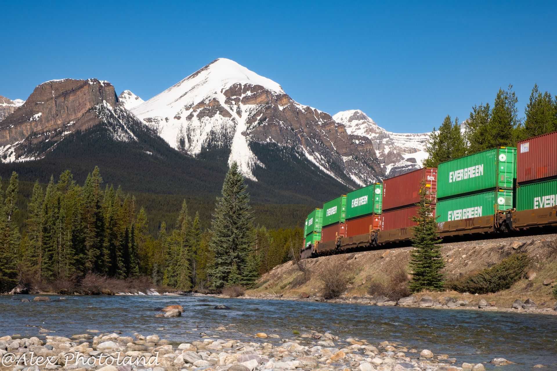 Train with cargo containers travels by a river with snow-capped mountains in the background. Green and red containers.