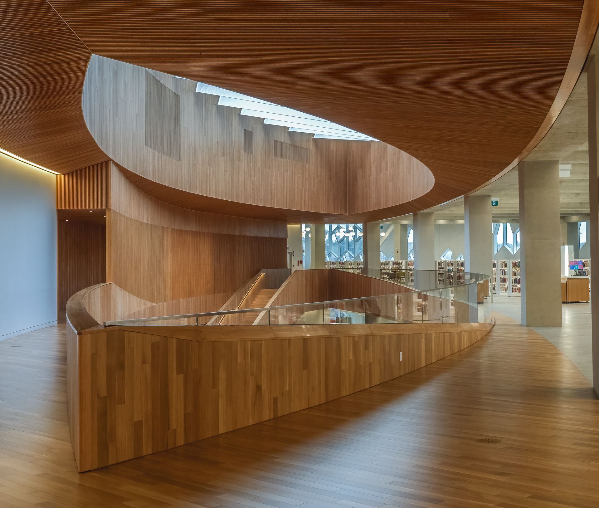 Wooden staircase spiraling up in a modern library. Natural wood tones throughout; curved walls, glass railing.