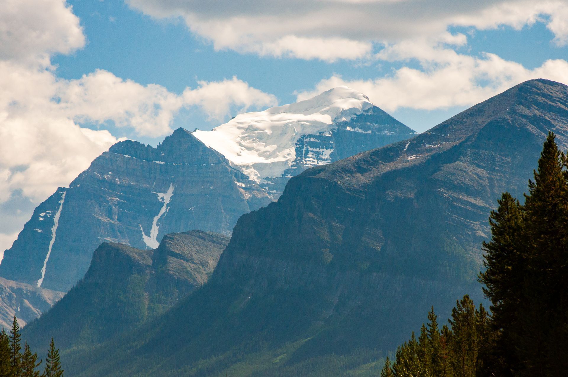 Snow-capped mountain peaks rising above forested slopes under a partly cloudy sky.