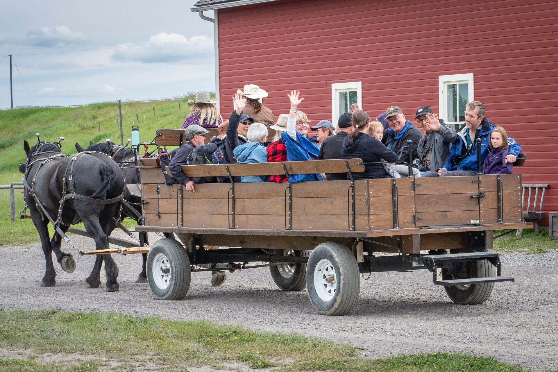 Horse-drawn wagon with people waving, near a red building and green grass.