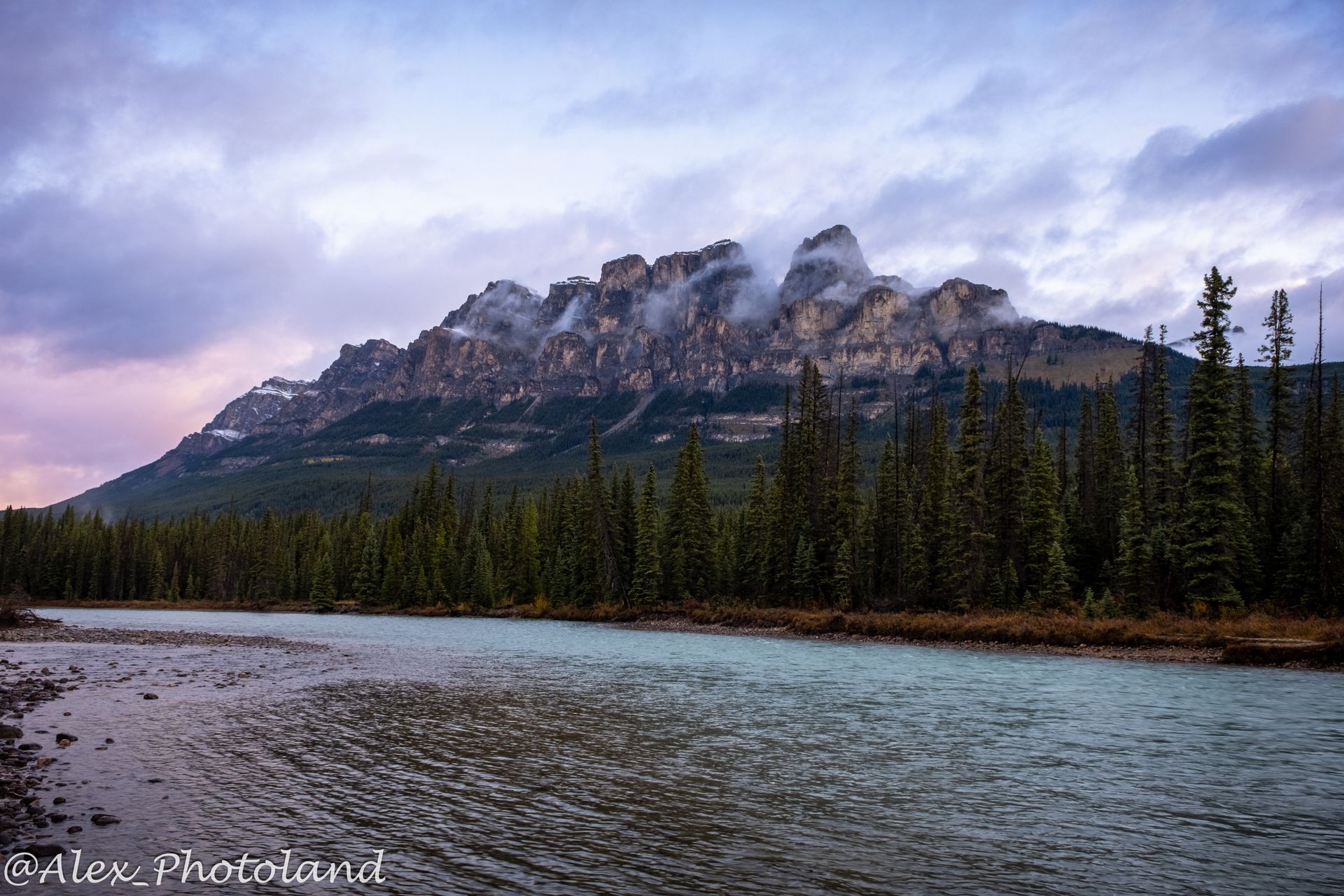 Castle Mountain, Canadian Rockies, with river in foreground and forested shoreline under a cloudy sky.