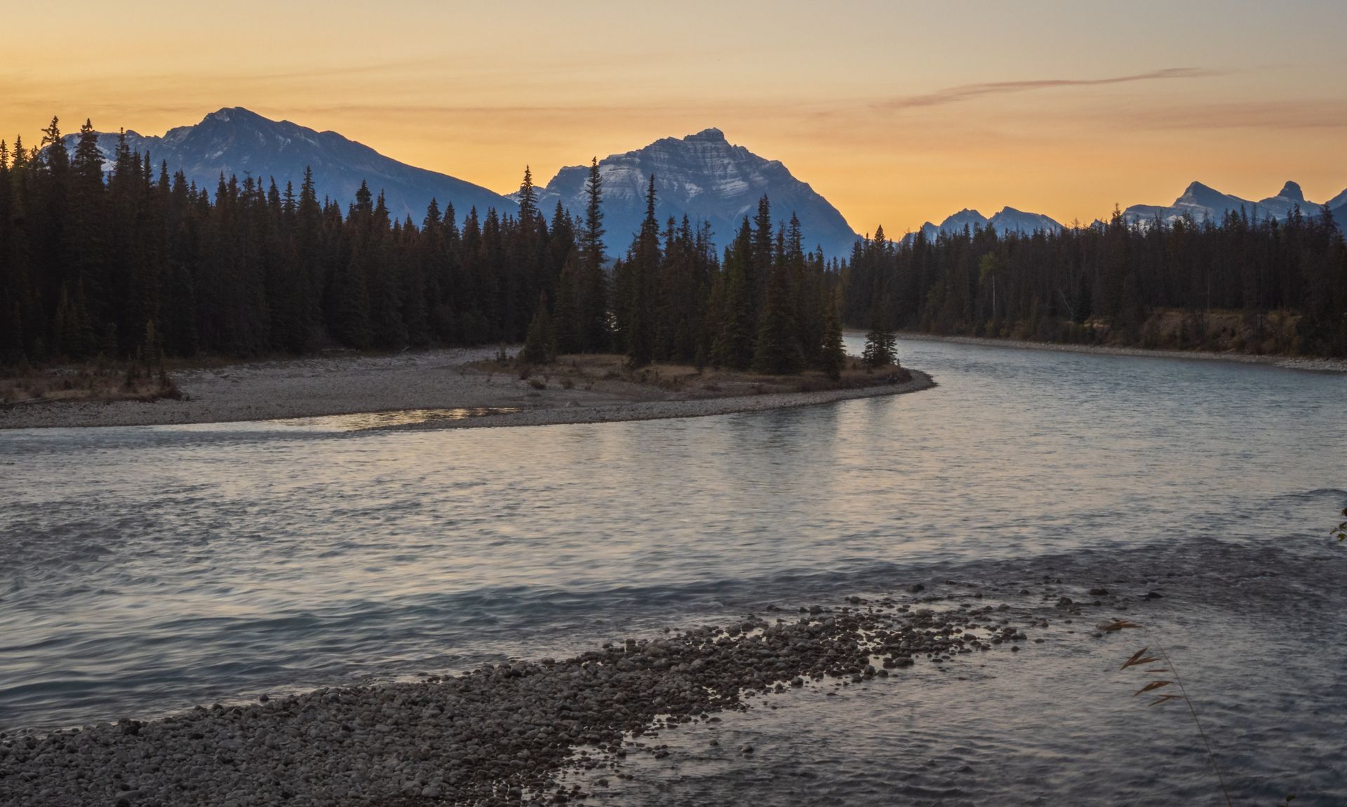 River flowing through a forest with mountains in the background at dusk; orange sky.