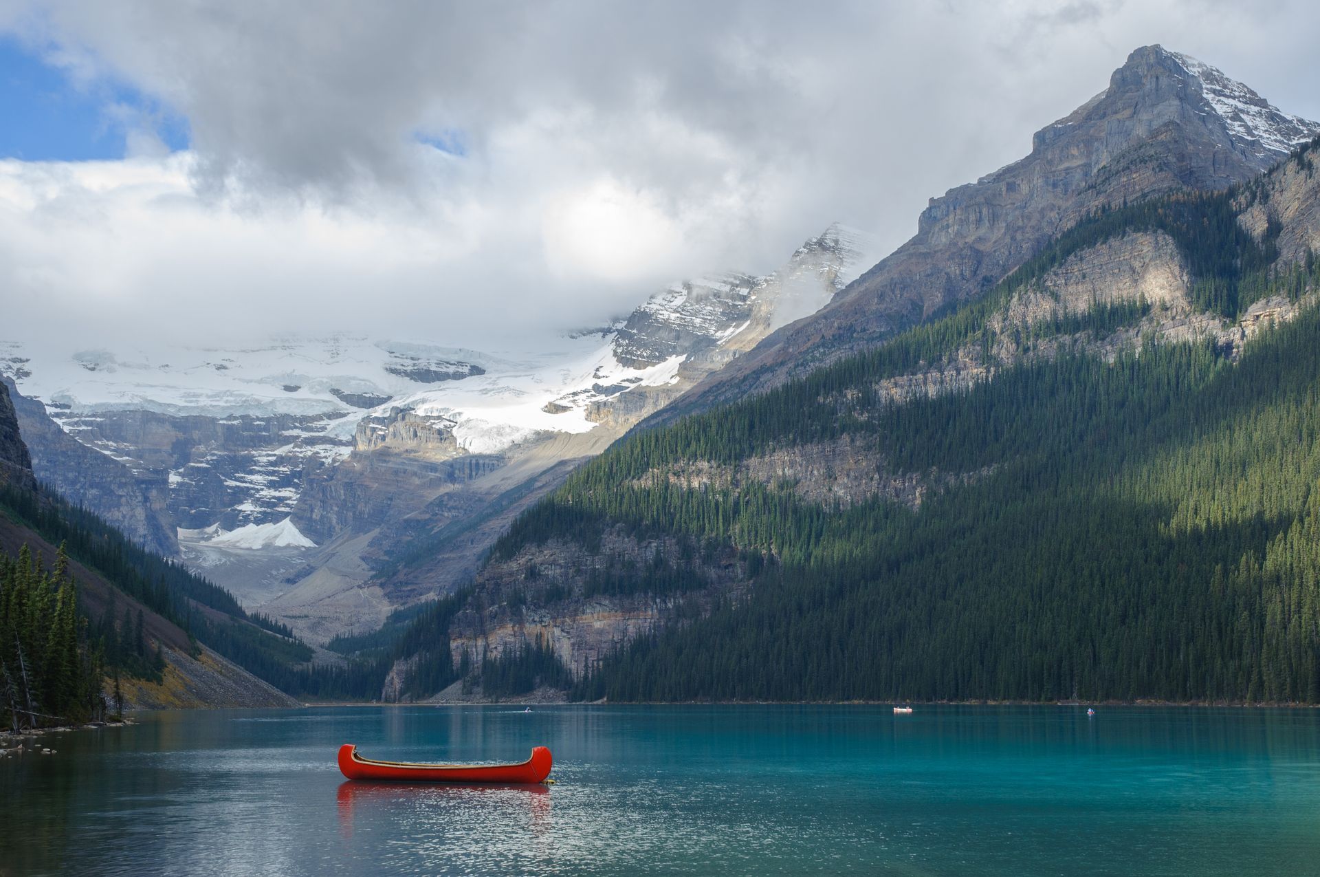 Red canoe on turquoise lake, framed by mountains, one snow-capped, in a scenic landscape.