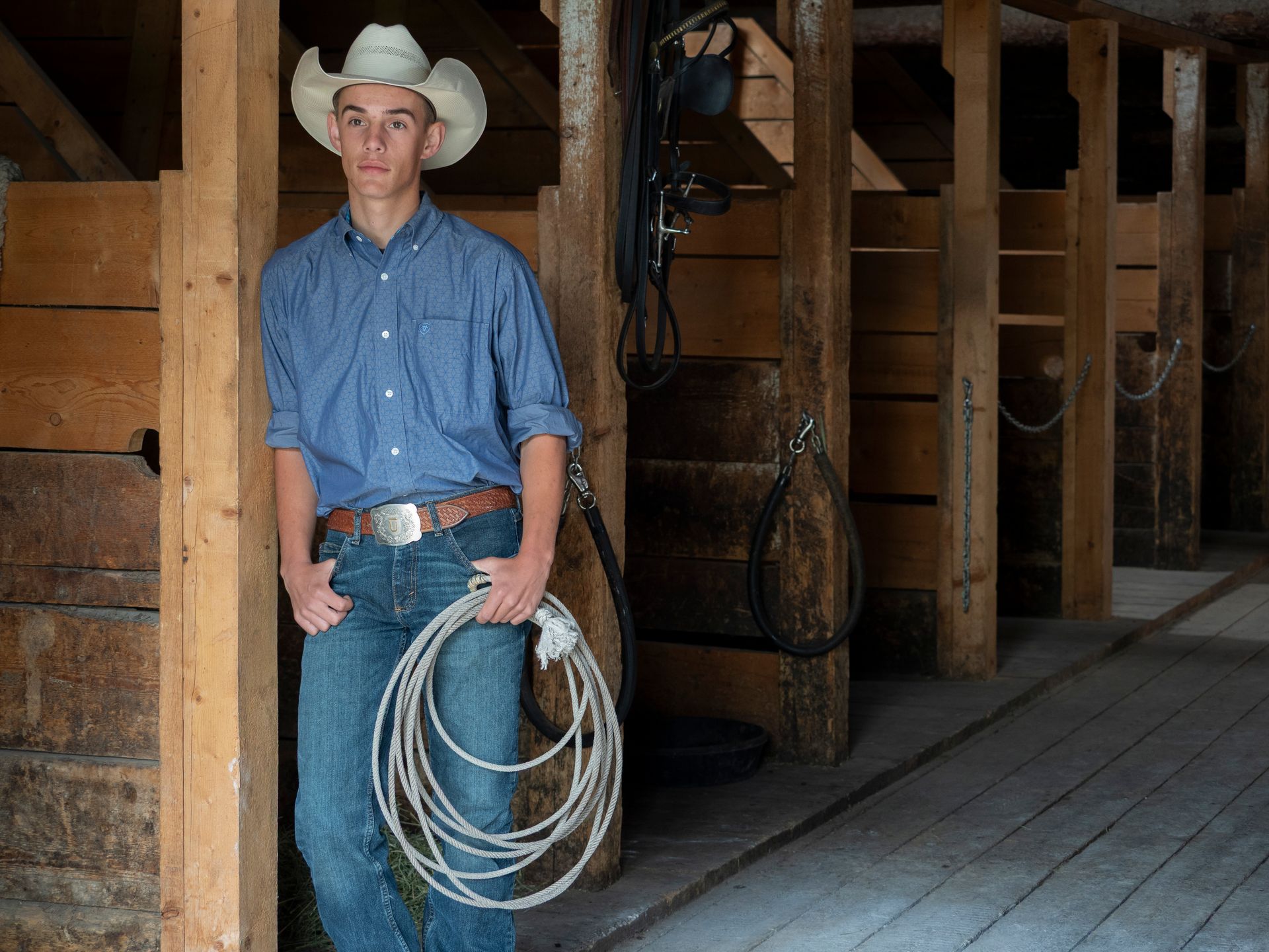 Cowboy in a stable, wearing a hat, blue shirt, jeans, holding a rope, leans against wooden posts.