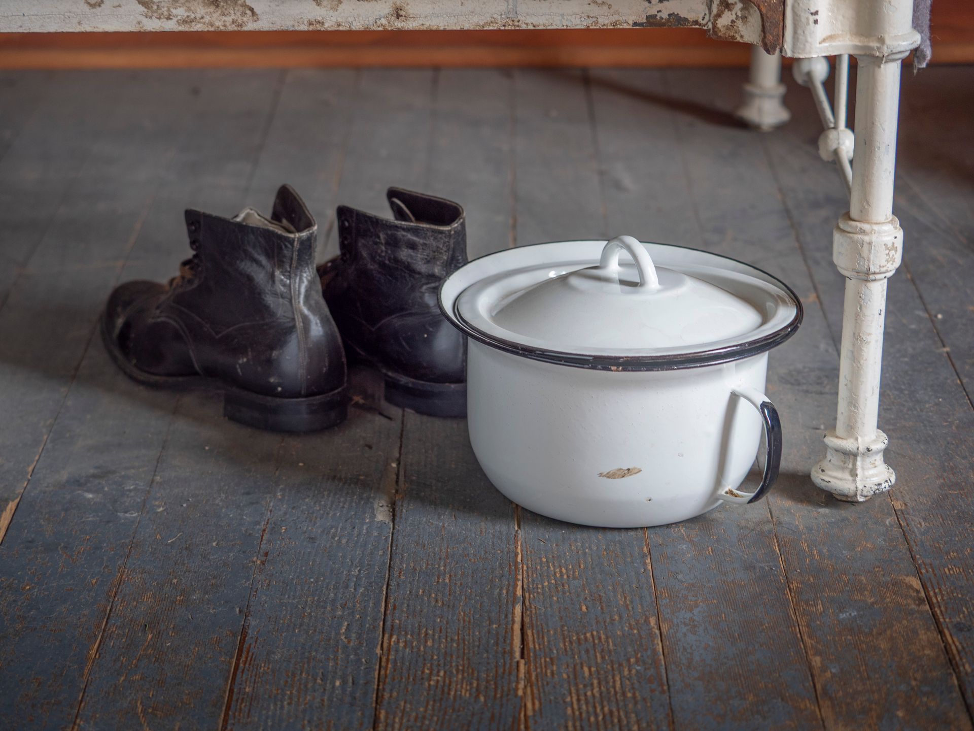 Black boots, white chamber pot with lid sit on weathered wooden floor beneath a bed.