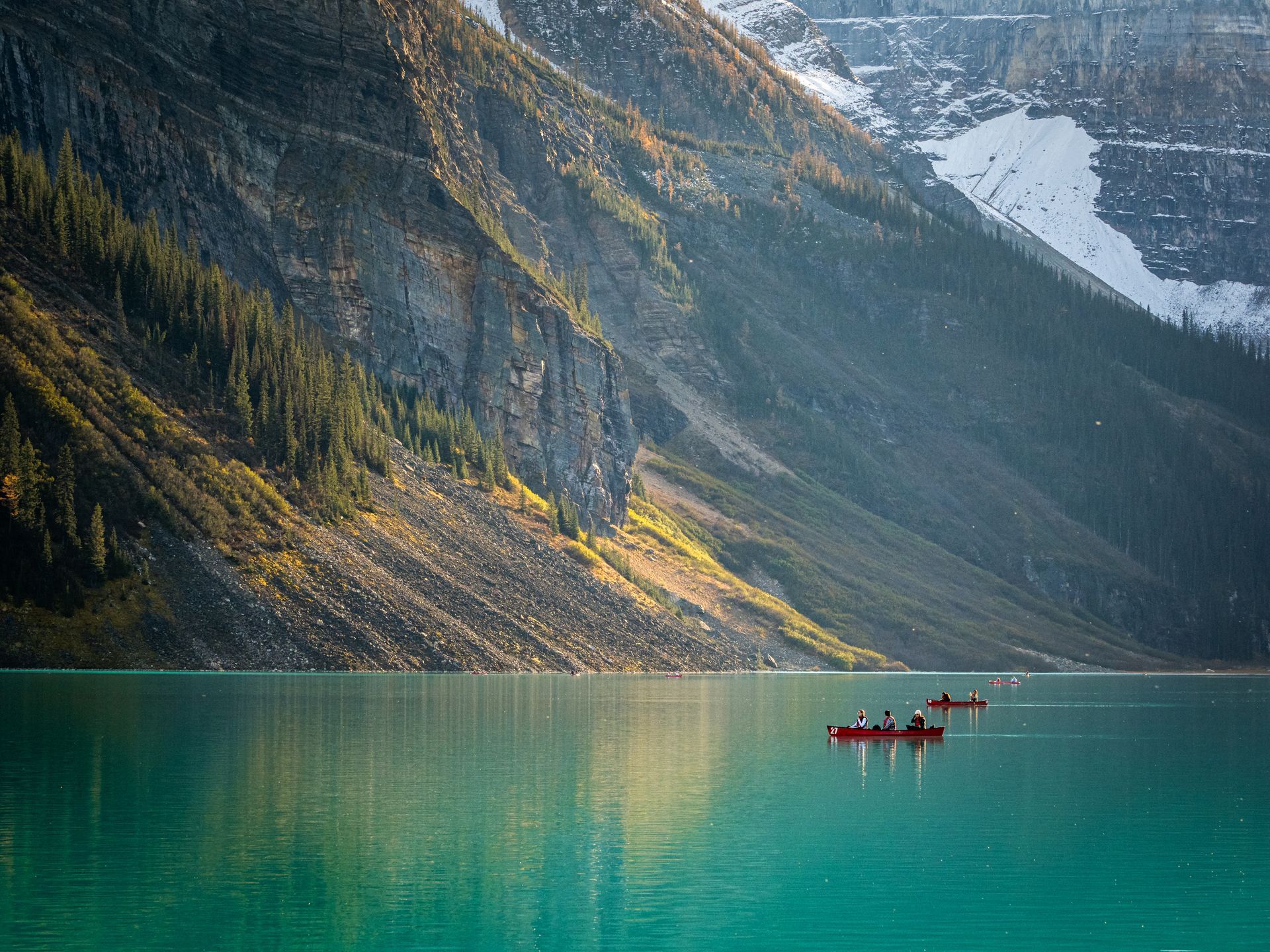 Turquoise lake with red canoes, beneath a rocky mountain with patches of snow.