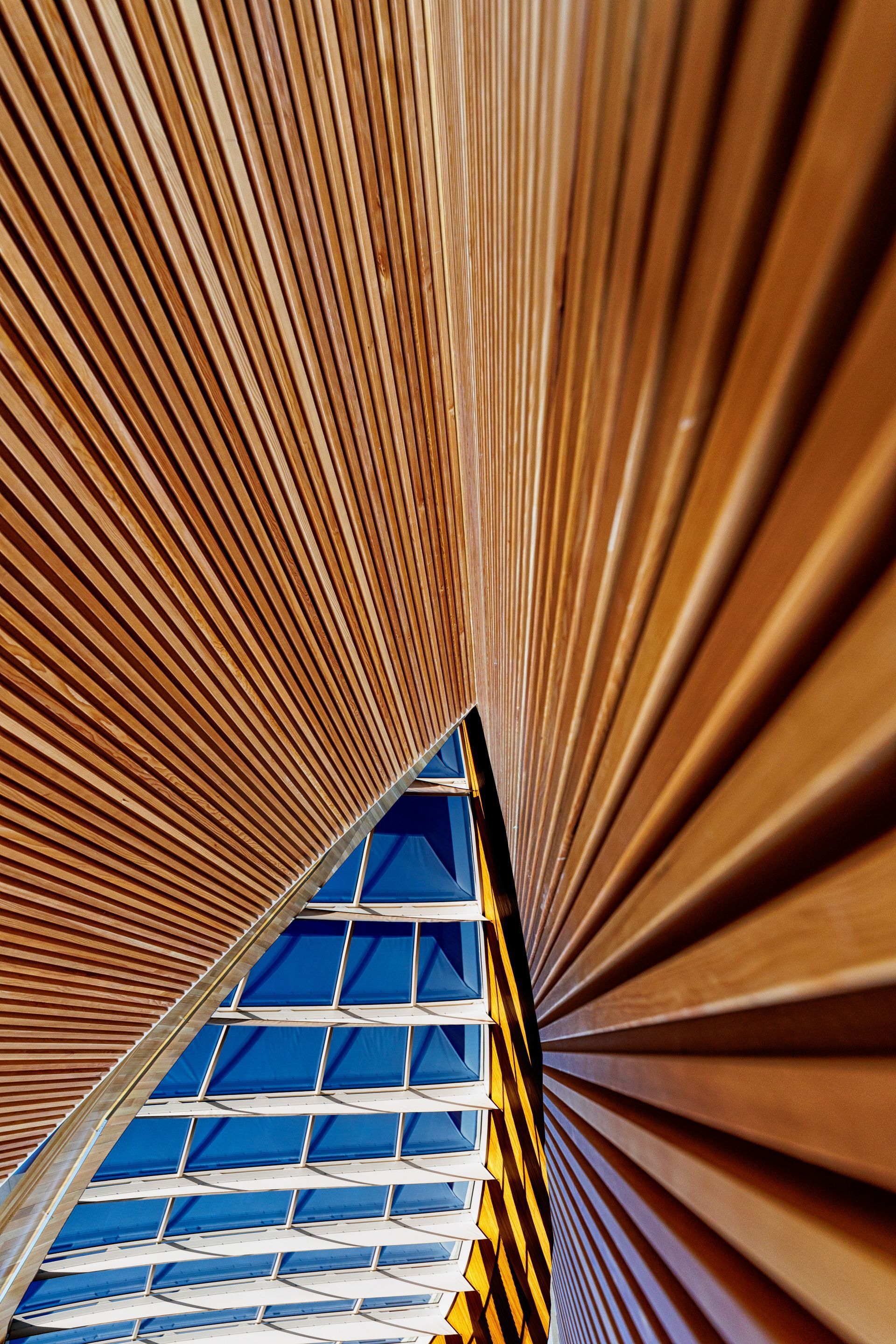 Wooden ceiling panels converging, revealing blue sky and glass structure.