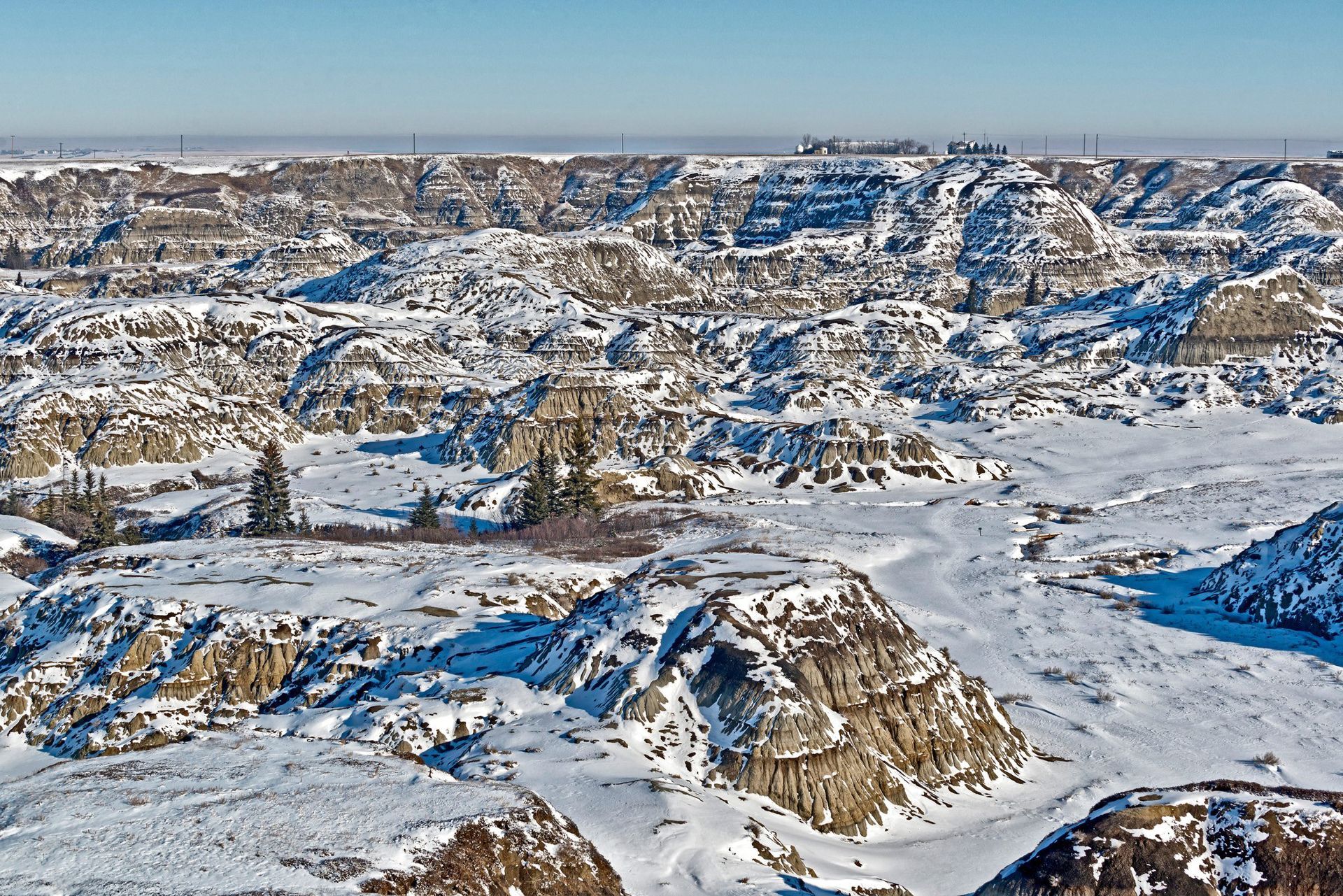 Snow-covered badlands landscape with layered rock formations under a clear blue sky.