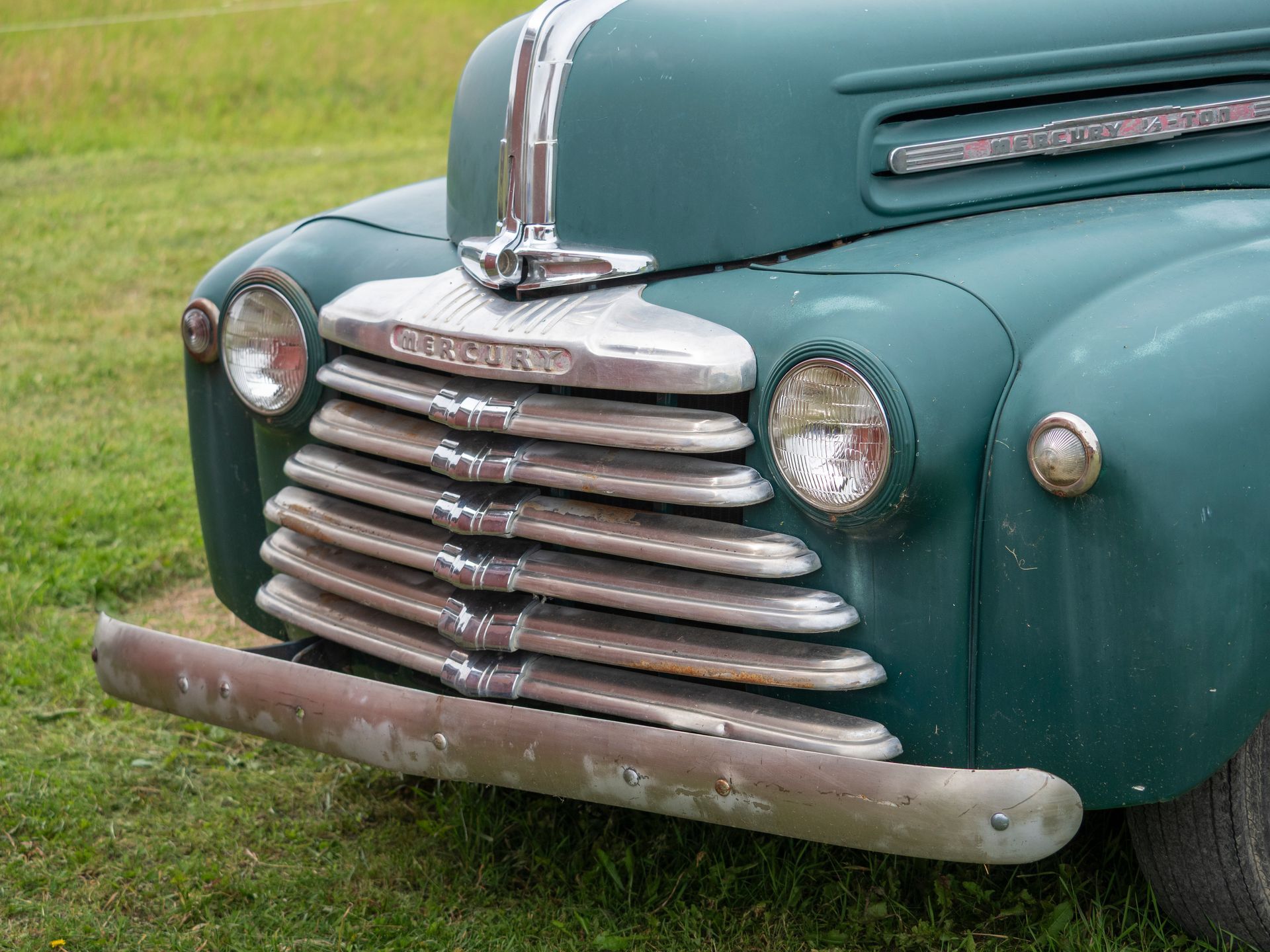 Green vintage Chevrolet truck's front with chrome grill and bumper, set on green grass.