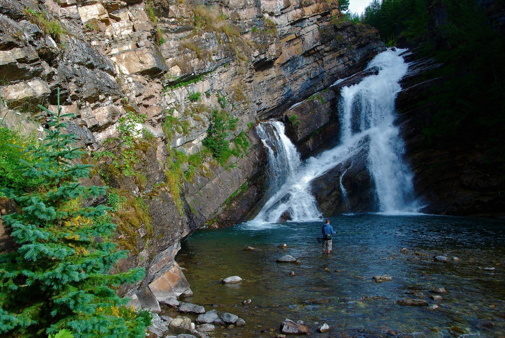 Waterfall cascading into a pool. Person stands in water; rocky cliff and trees frame the scene.