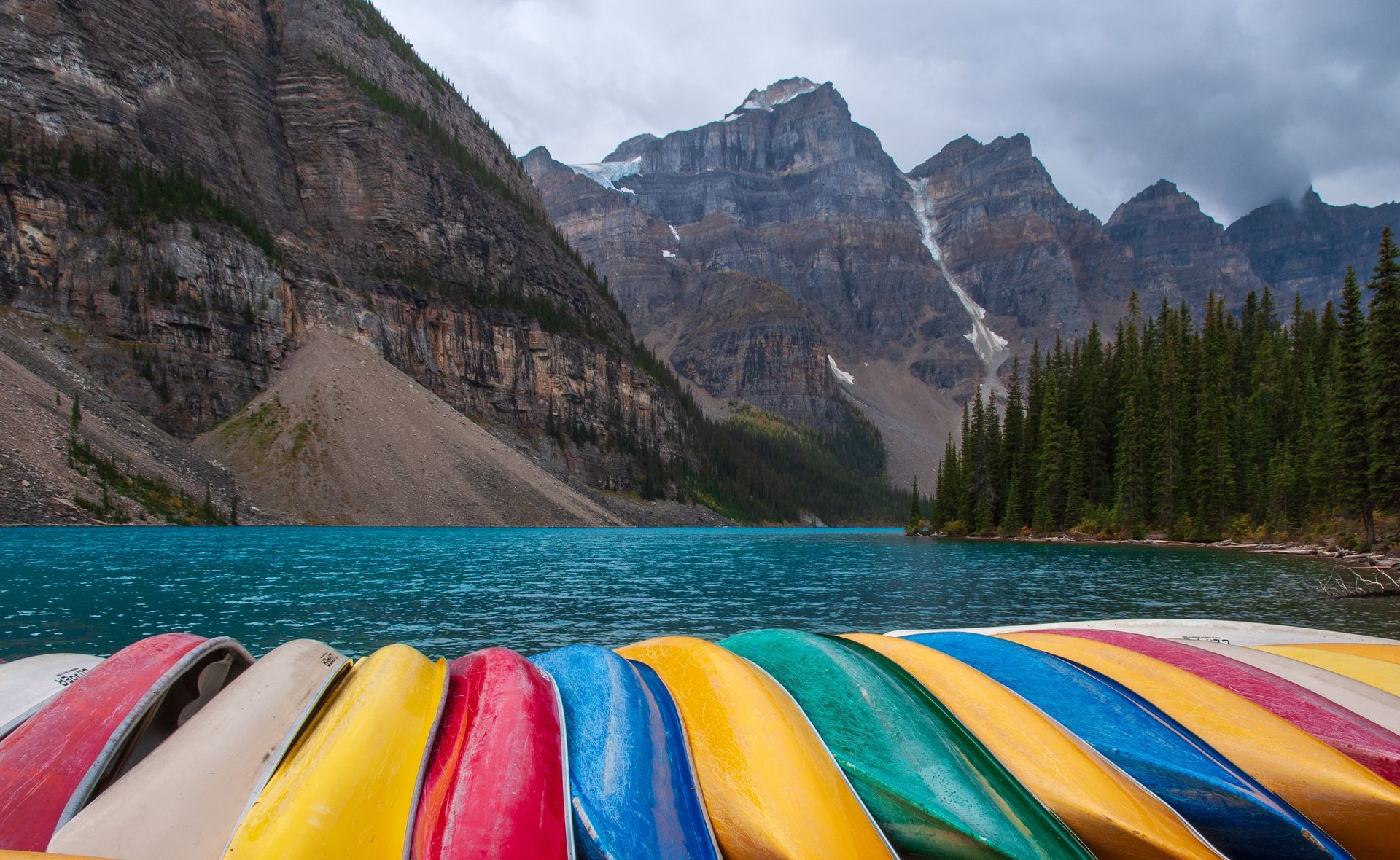 Colorful canoes line the shore of a turquoise lake with mountains in the background.