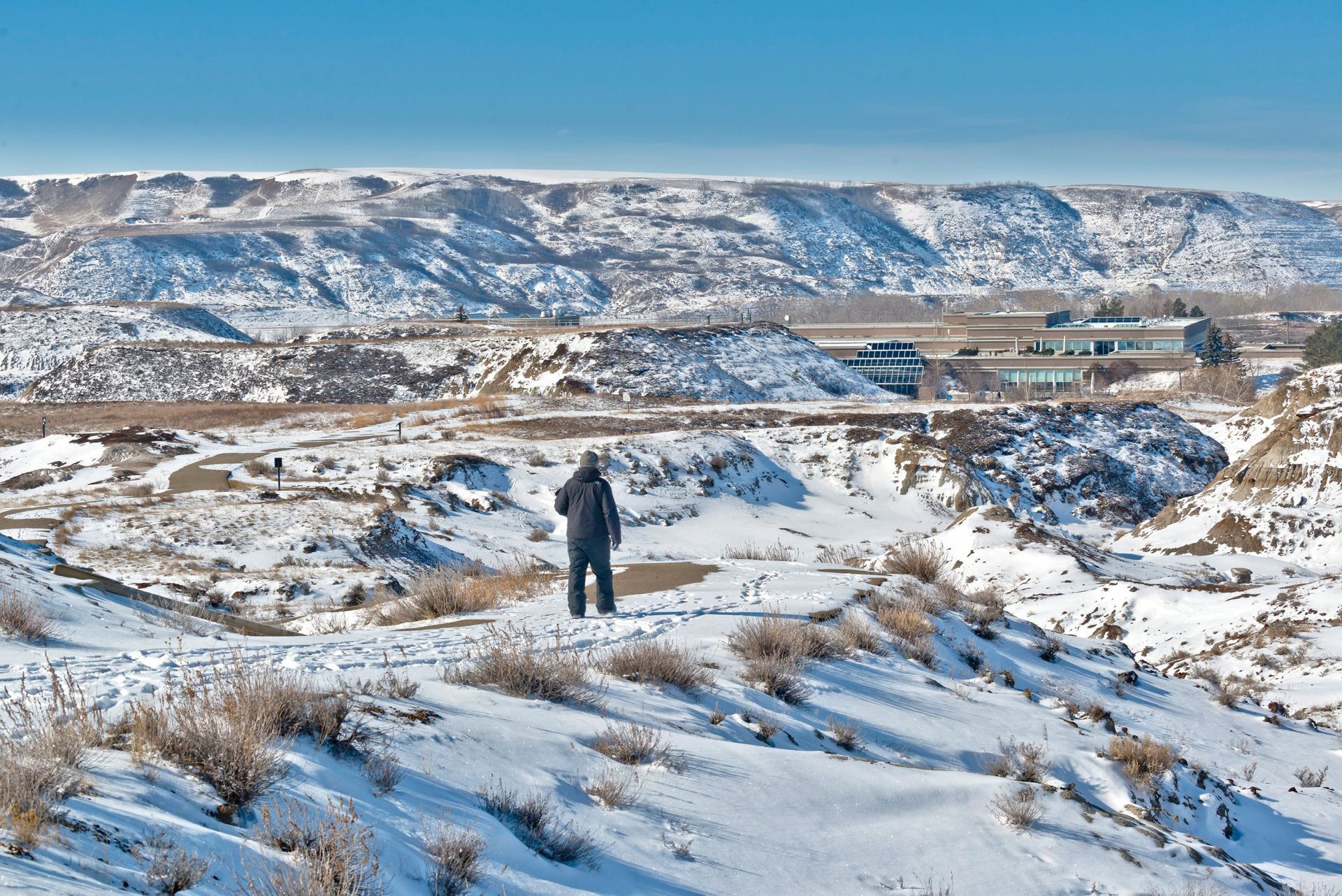 Person standing in snowy landscape, overlooking buildings and distant hills under a bright sky.