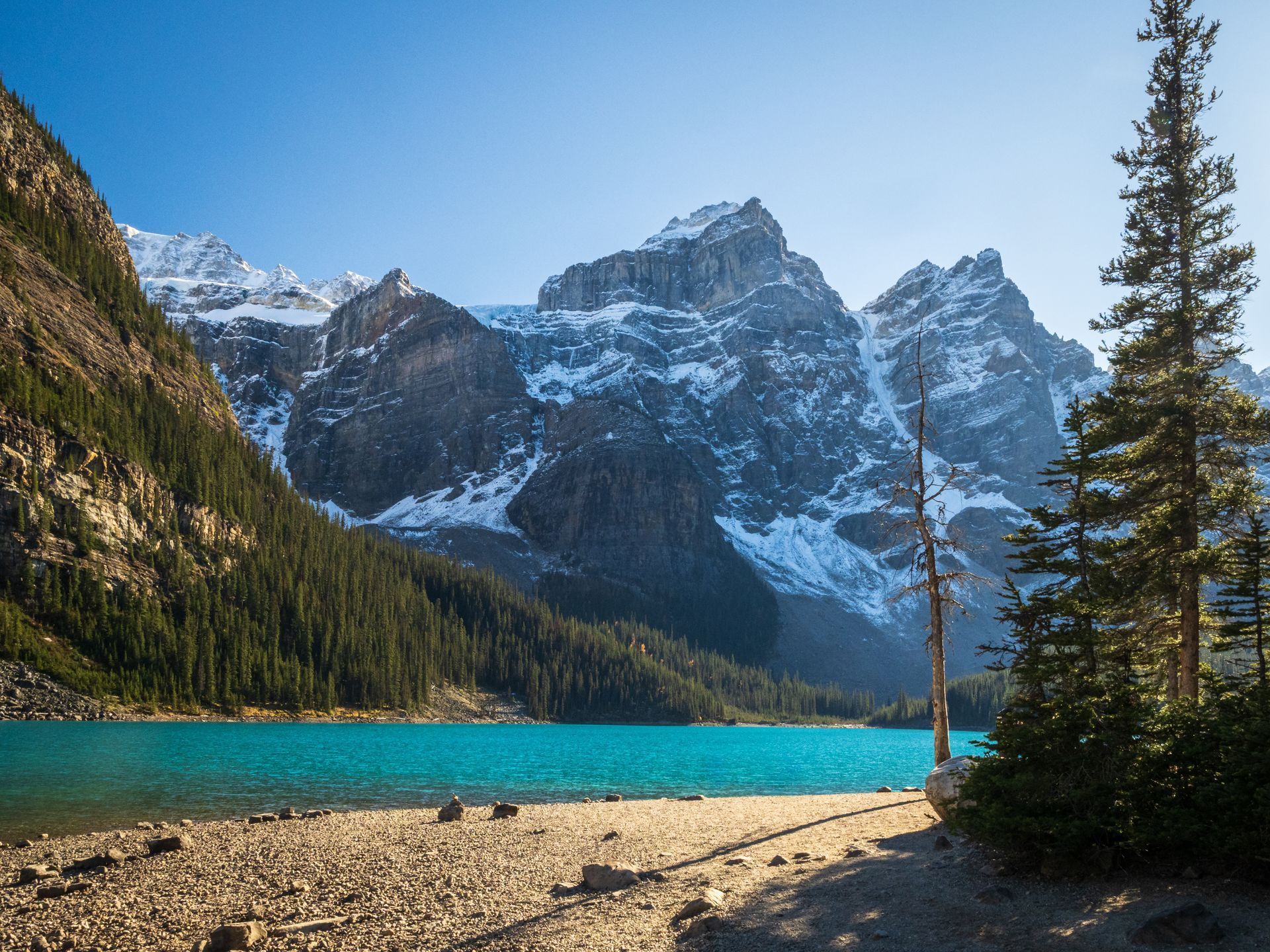 Turquoise lake with snow-capped mountains and evergreen trees under a blue sky.