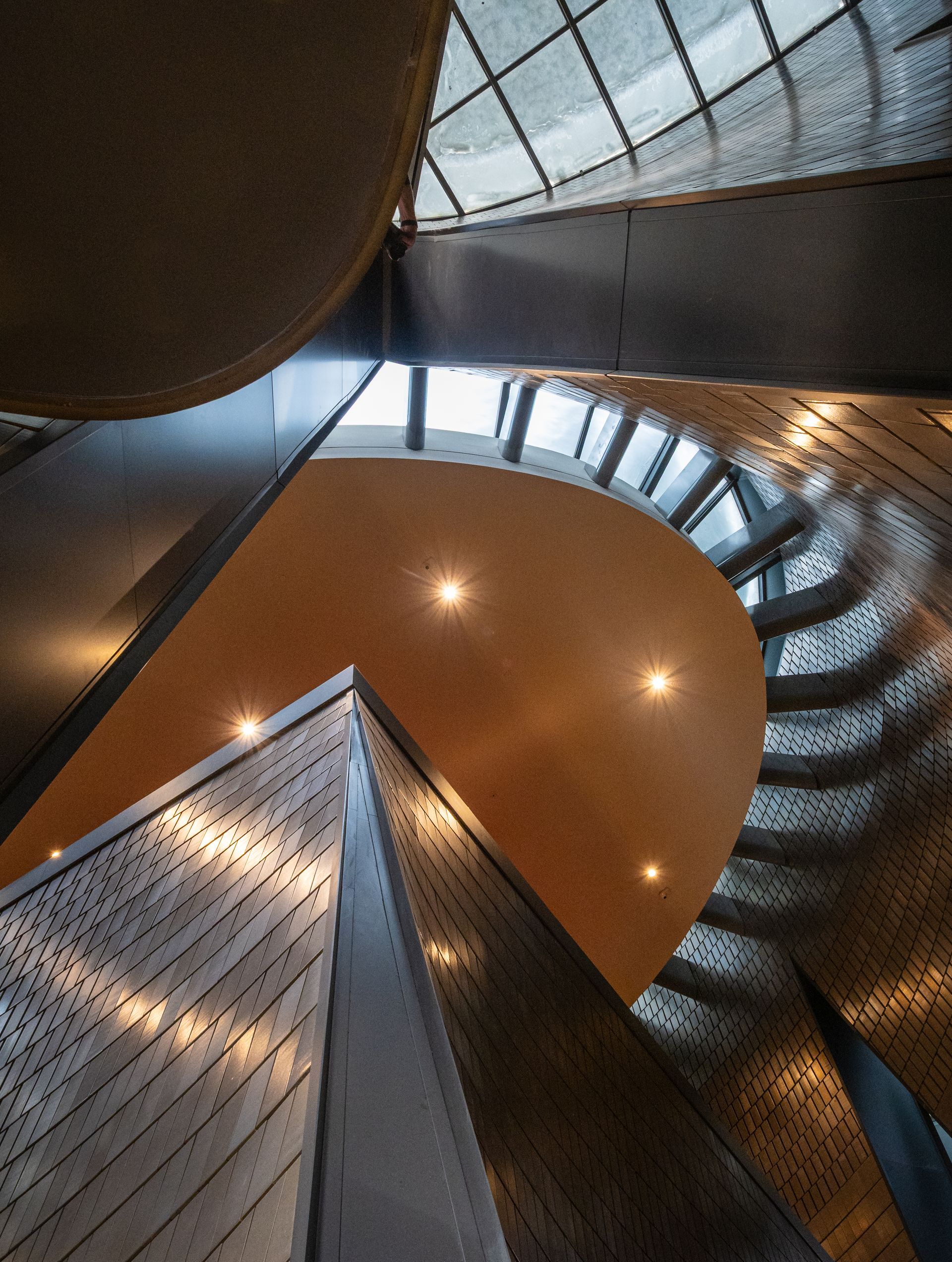Looking up a modern, spiral staircase with gold and silver accents and recessed lights.