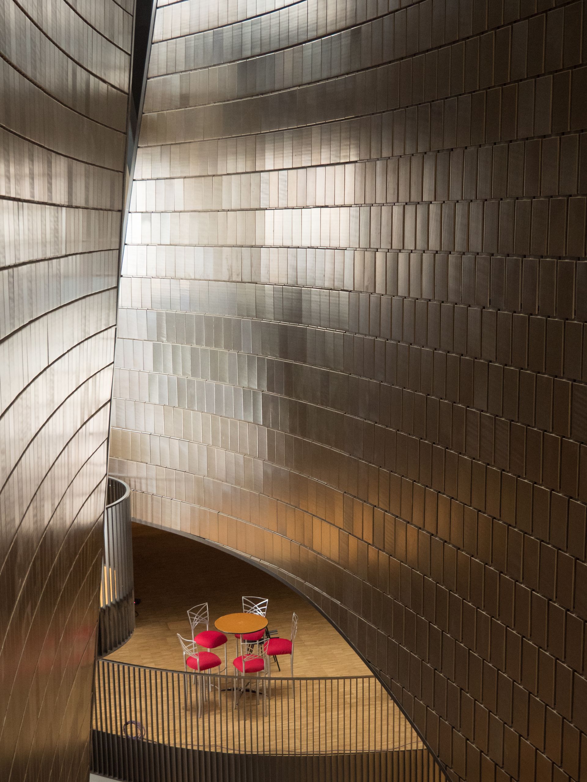 Curved, metallic-tiled walls enclose a small seating area with red chairs and a yellow table.