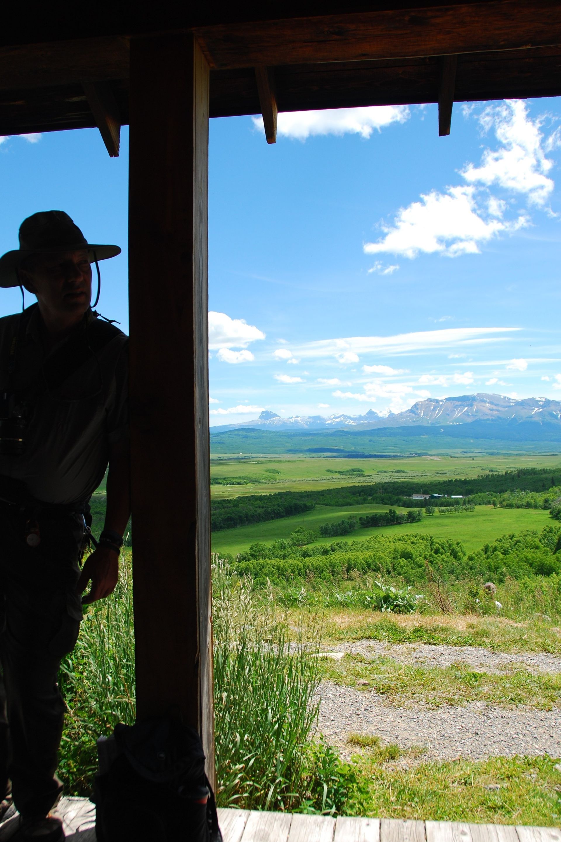 Person in shadow on porch, overlooking green valley and mountains under a blue sky with clouds.