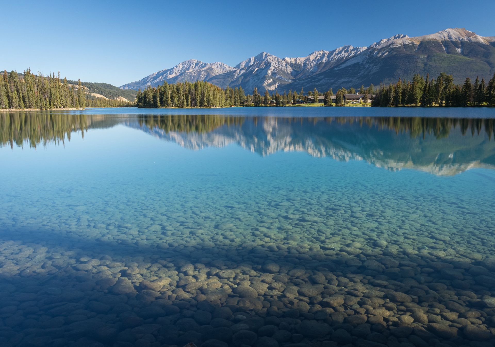Clear turquoise lake with rocky bottom reflecting mountains and trees on a sunny day.