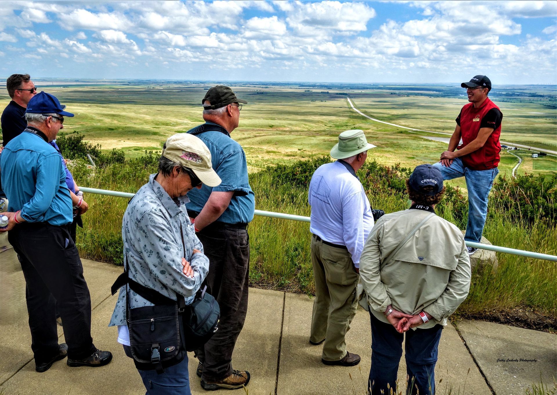 People on a scenic overlook listen to a man in a red shirt. Green fields and blue sky in the background.