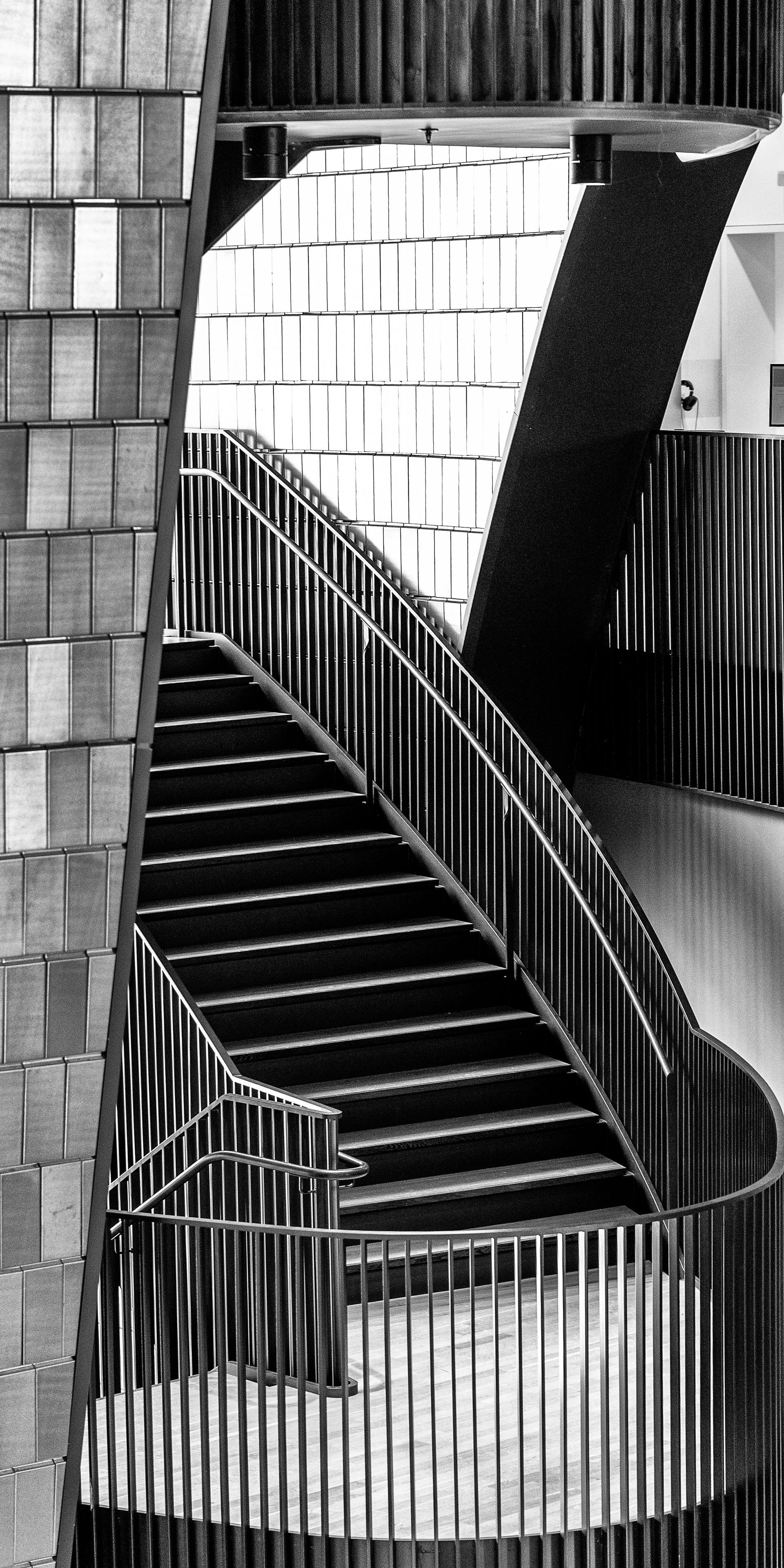 Black and white photo of a staircase with metal railings. Brick wall on the left and geometric patterns in the background.
