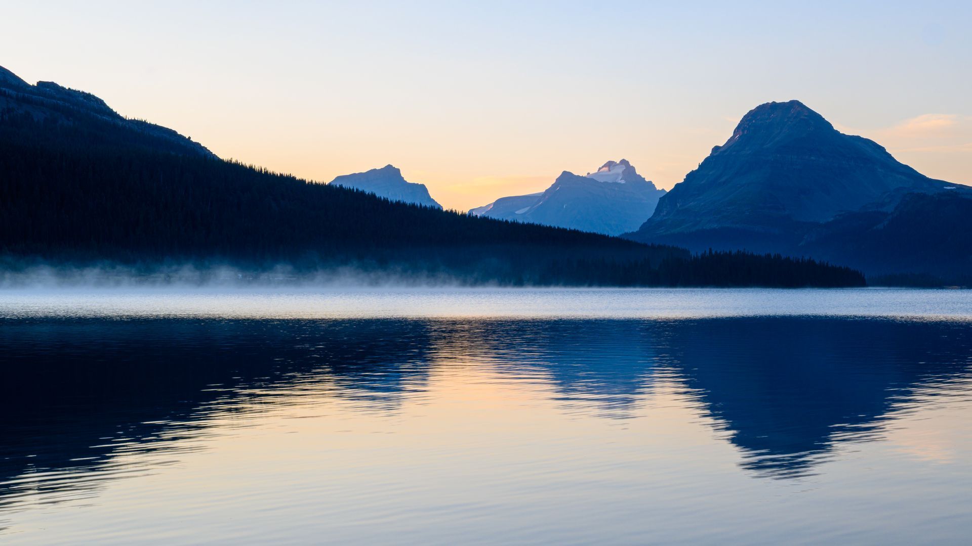 Mountains reflected in a still lake; misty atmosphere with blue and gold hues.