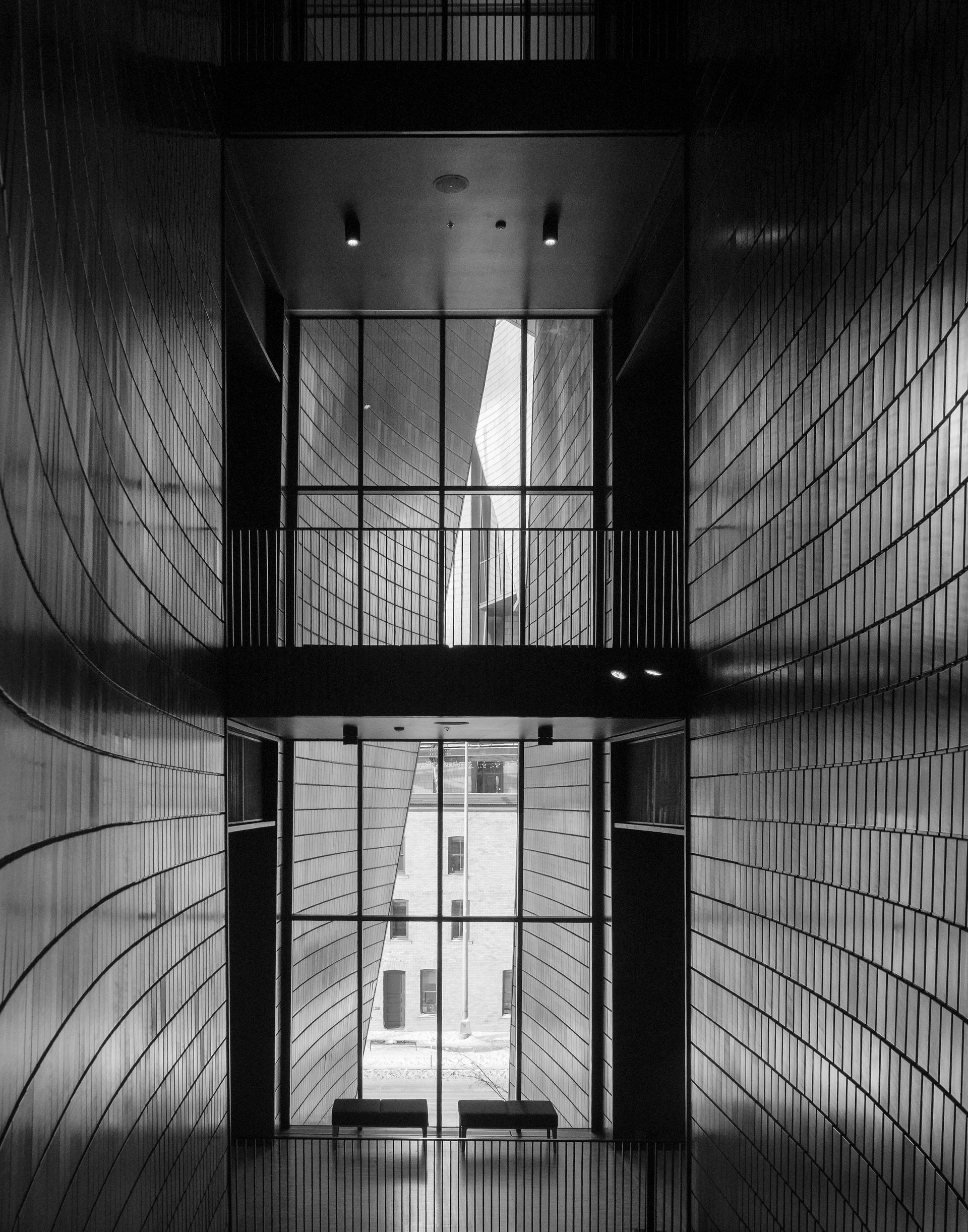 Black and white interior shot of a long hallway with textured walls leading to exterior windows. Two benches are visible.