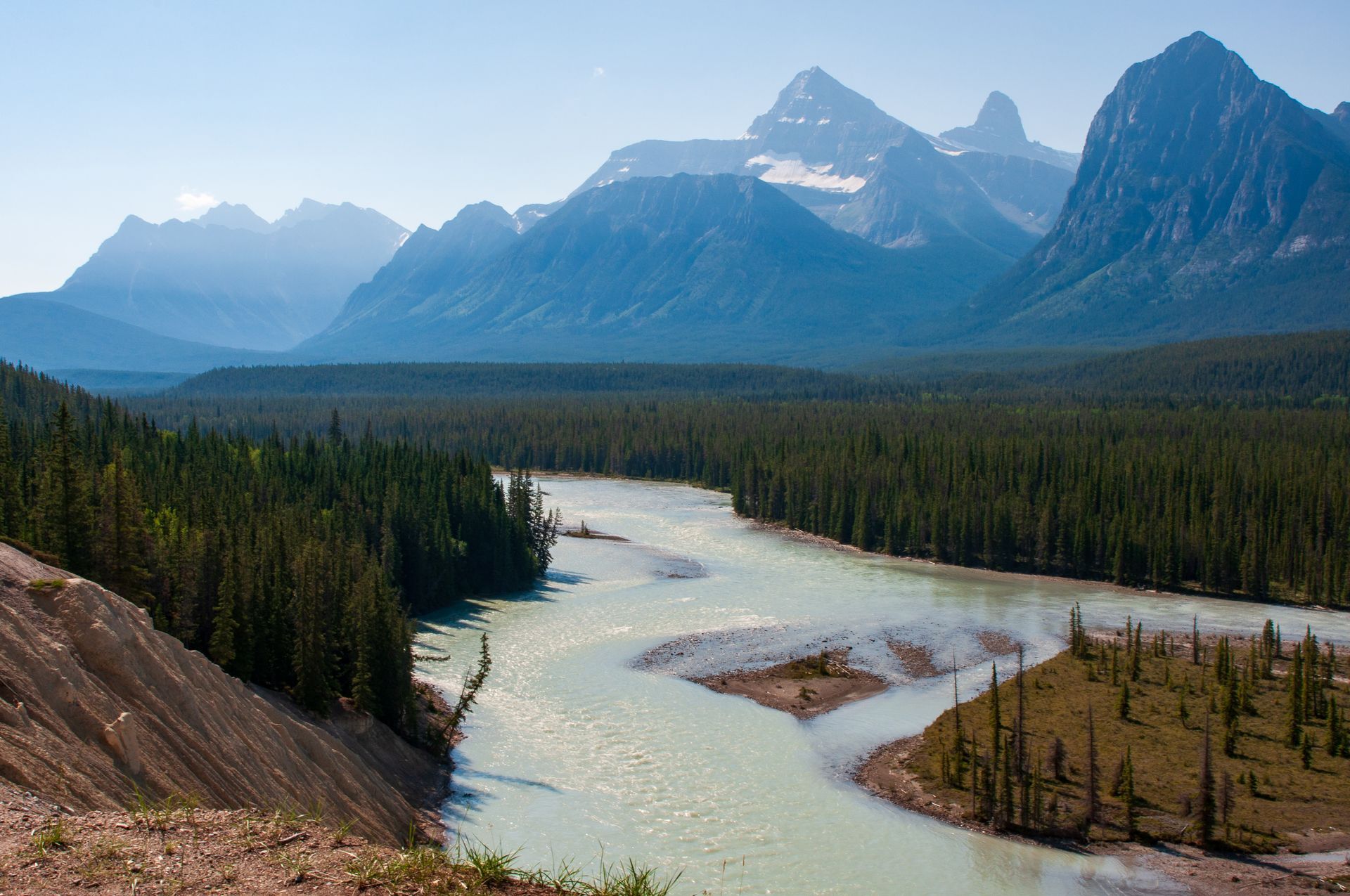 River winding through a dense forest with mountains in the background, under a bright blue sky.