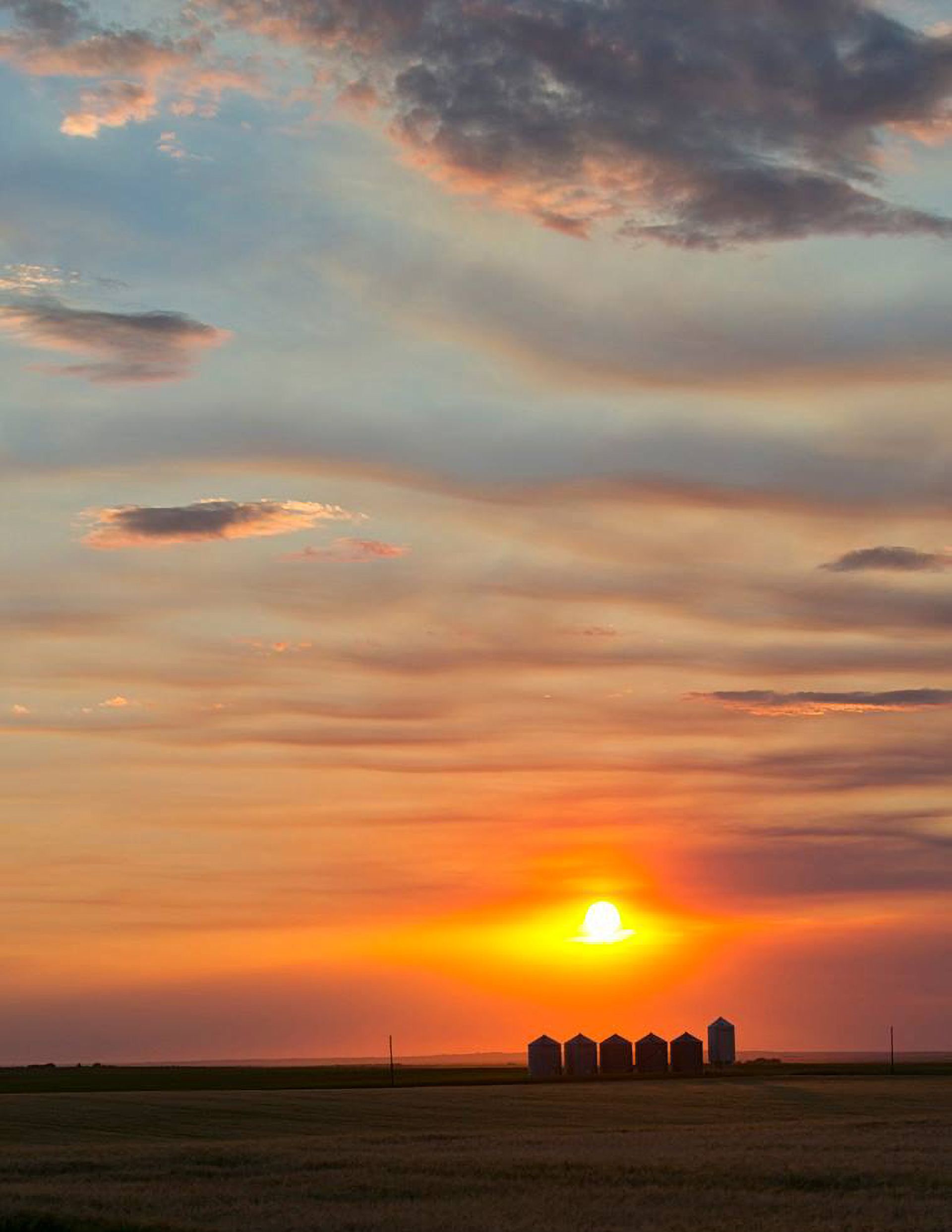 Sunset over silos; orange and yellow sky with wispy clouds; fields in the foreground.