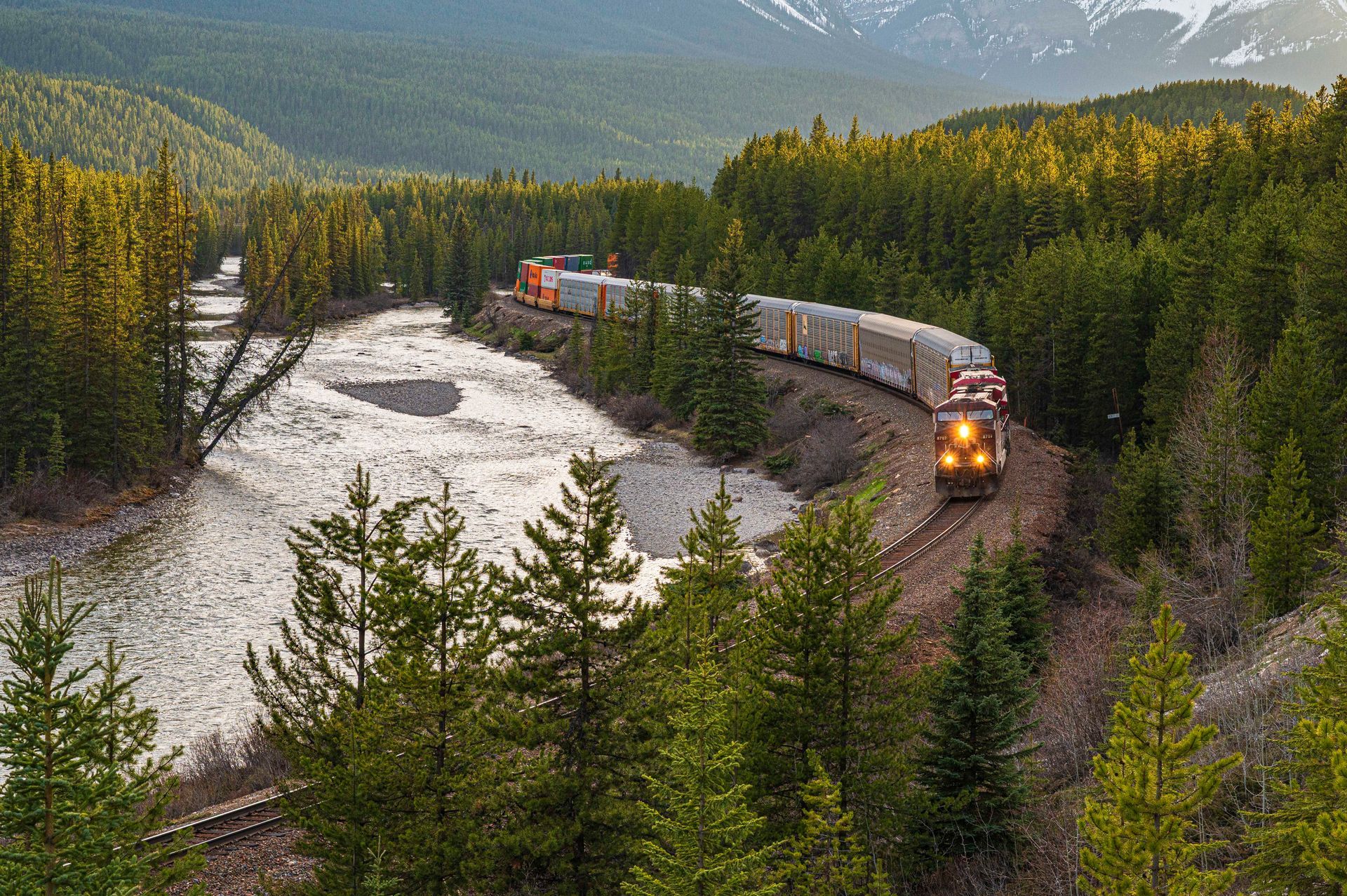 Train winding through a forest along a river with snow-capped mountains in the background.