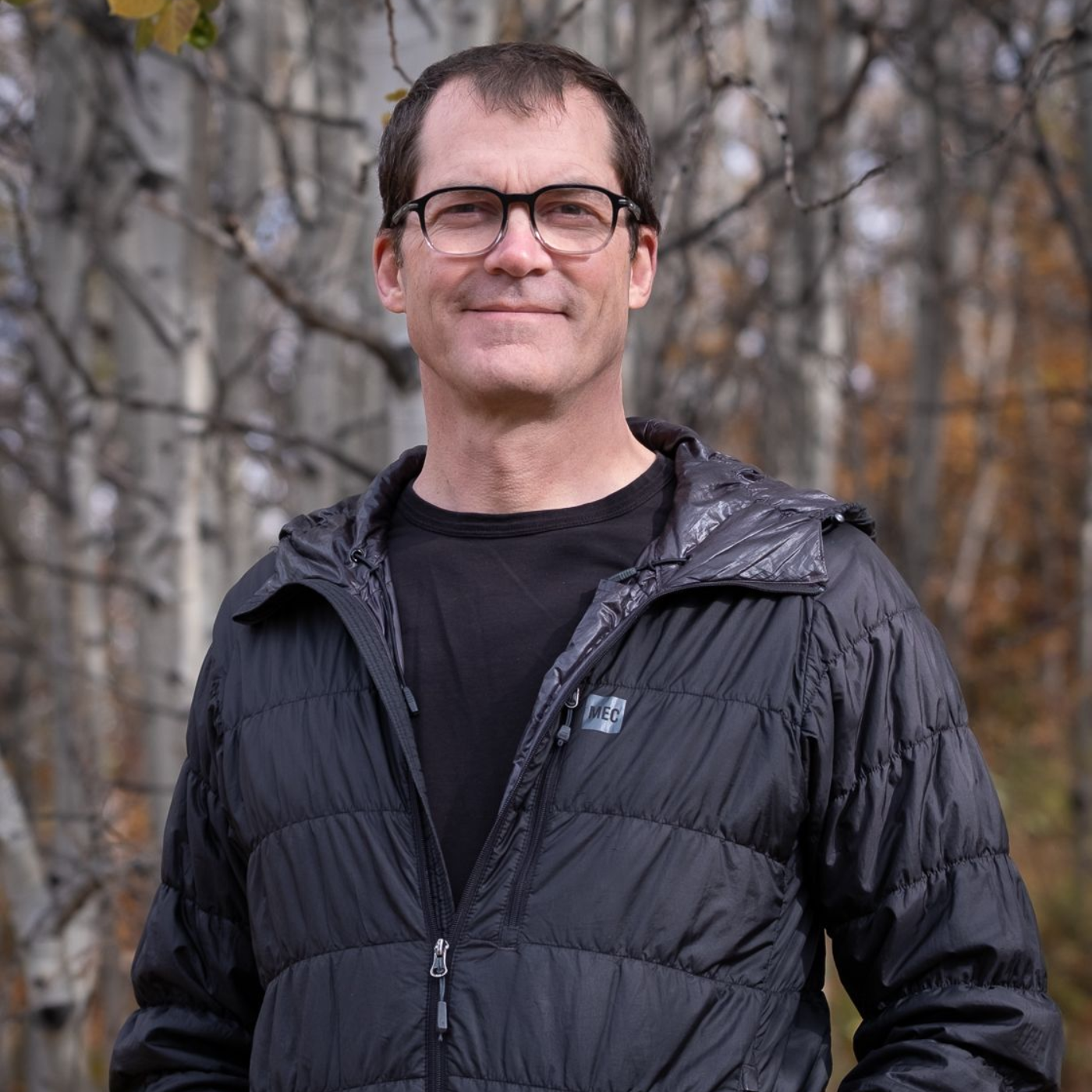 Man with glasses and black jacket stands outside near trees.